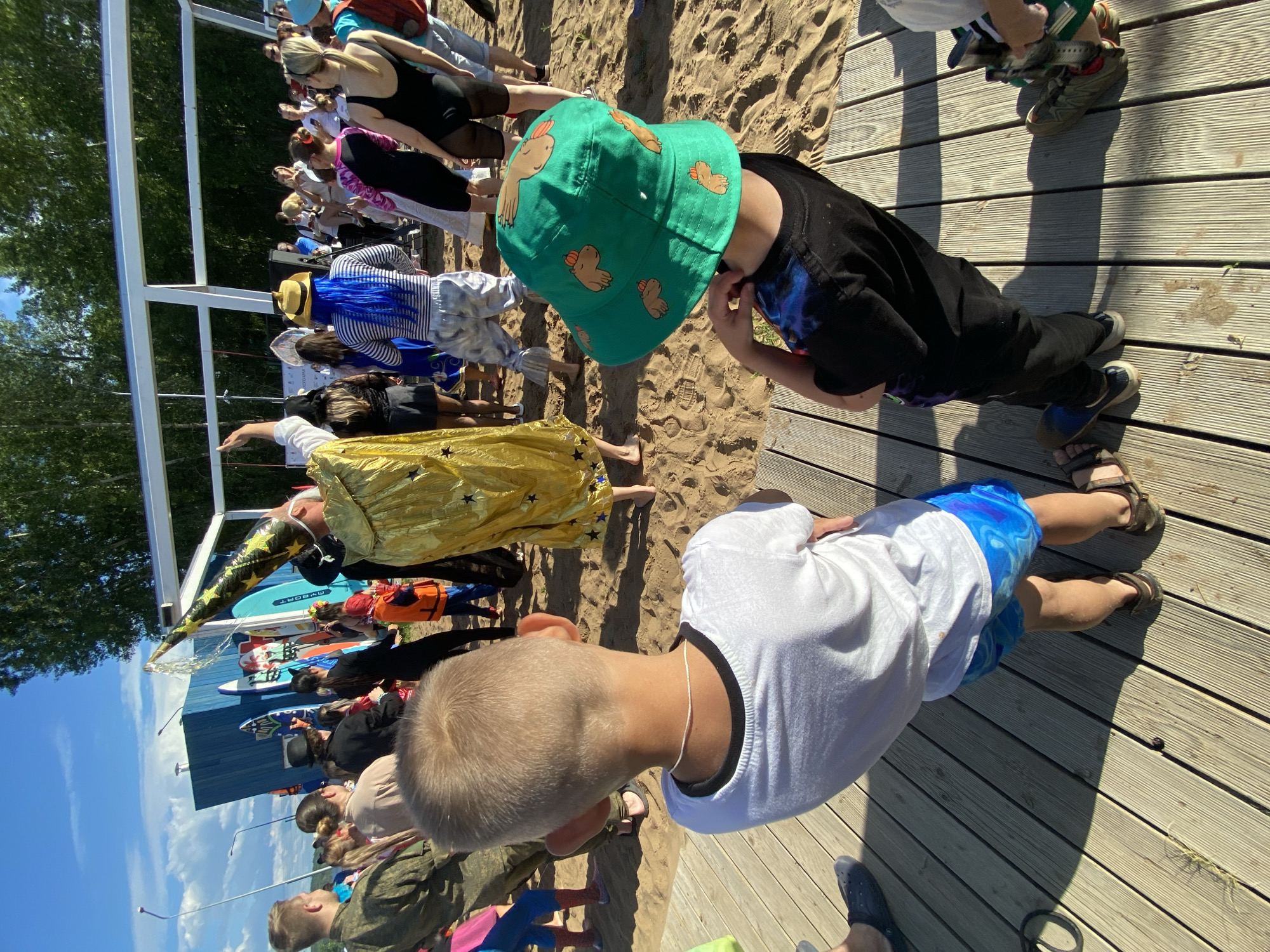 The image shows an outdoor event on a sandy beach with a wooden boardwalk. Several people are gathered, some in costumes. In the foreground, two young boys are visible from behind, facing away from the camera. The boy on the left wears a white t-shirt and blue patterned swim trunks. The boy on the right wears a black t-shirt and a green bucket hat with cartoon capybara illustrations. Behind them, a person in a golden, star-patterned cape and conical hat appears to be pouring liquid from the hat. Other attendees are dressed in various outfits, including a person with bright blue hair and a striped shirt. The background features a blue structure with surfboards leaning against it, trees, and a bright blue sky with some clouds. The location is identified as Chaikovsky, Russia.