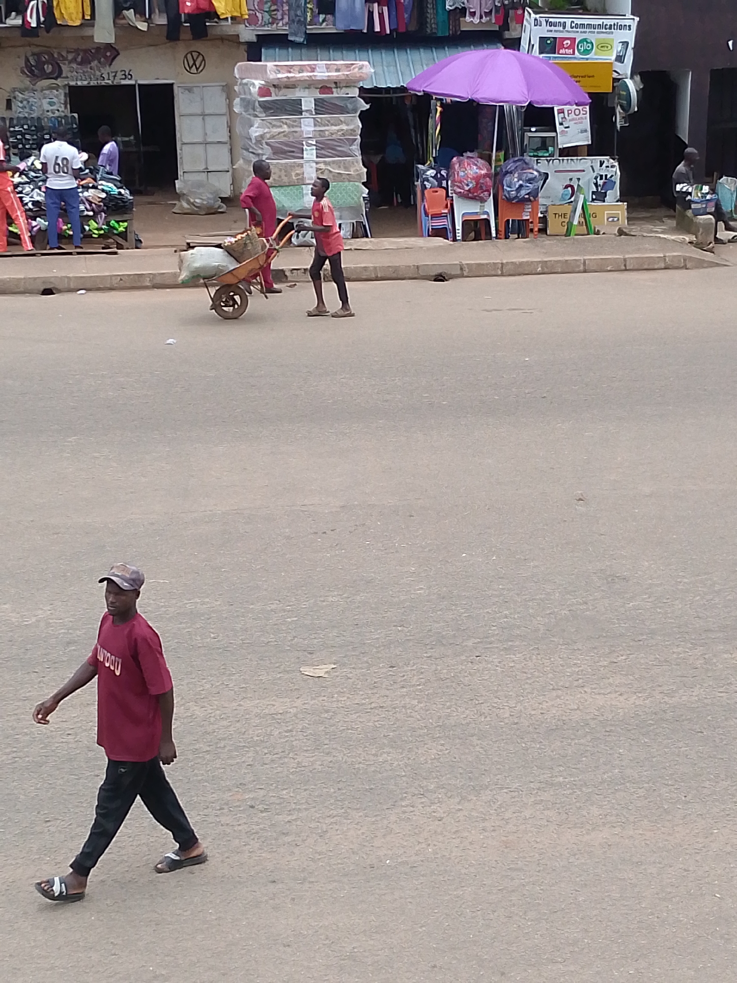 In an outdoor, urban environment, likely a street market, a person is walking towards the left side of the frame. They are wearing a maroon t-shirt with a white inscription and black pants, along with open-toed sandals. In the background, several other individuals are present. Two people are near a cart with goods, one pushing it and the other walking alongside. Another person in a white t-shirt with the number "68" is standing near a display of items. Shops with various goods, including mattresses, clothing, and electronics, line the street. A purple umbrella is set up in front of one of the stalls. The ground is paved, and a curb is visible.
