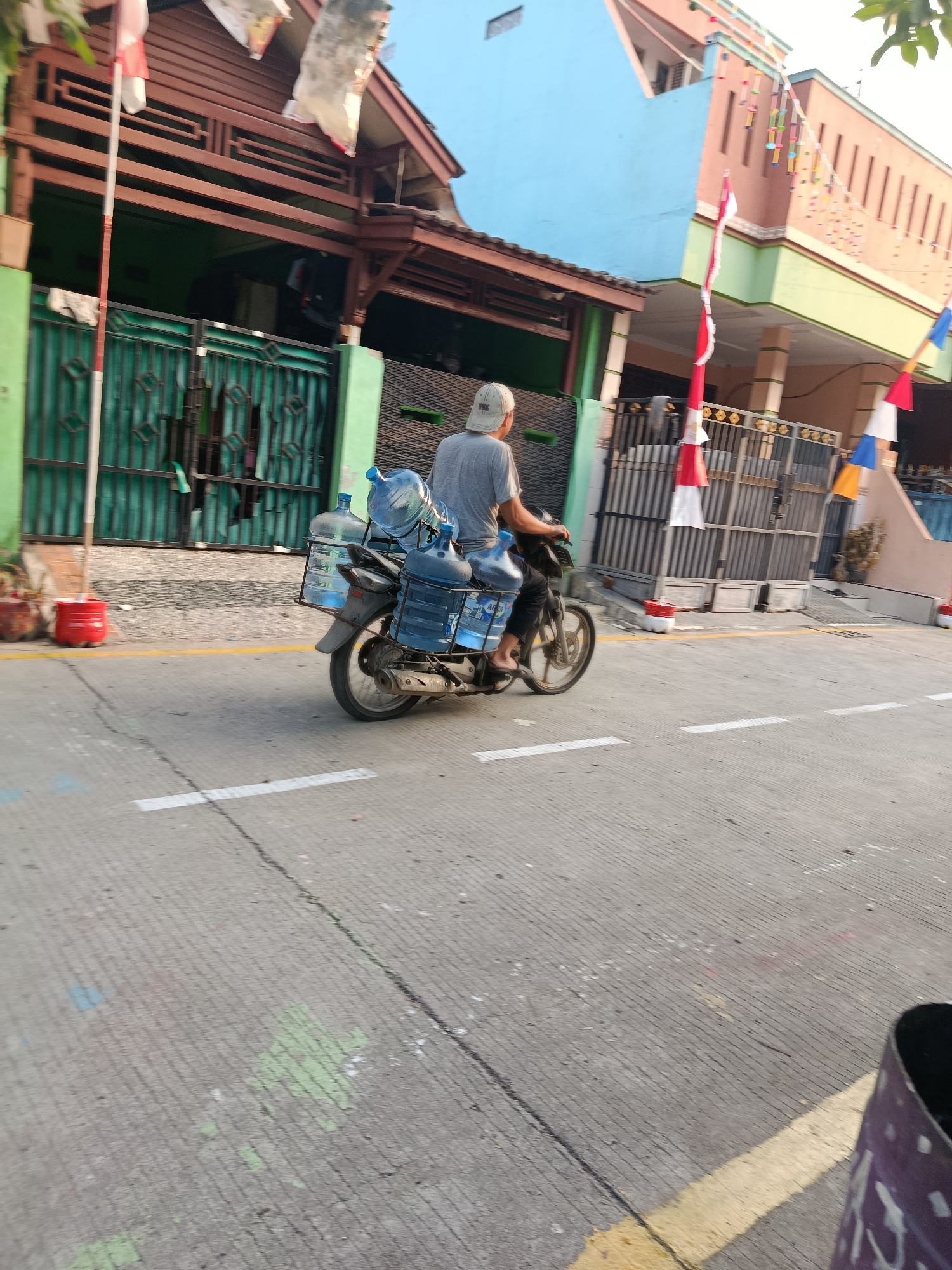 An Indonesian man wearing a grey t-shirt and a white baseball cap is riding a motorcycle on a paved road in Wanasari, Indonesia. The motorcycle is equipped with a rack on the back, carrying five large, clear plastic water jugs. The jugs are labeled with blue text, indicating they contain "AQUA" brand water. The man is positioned in the center of the frame, facing away from the camera and to the left. His legs are visible, and he appears to be wearing dark pants and sandals. The motorcycle is moving forward, creating motion blur.
The environment around the road includes residential buildings. To the left of the road, there is a green fence and the side of a building with a brown wooden facade. Red planters with some greenery are visible near the fence. To the right of the road, a multi-story building with blue and beige sections is prominent, decorated with colorful festive banners and flags. A grey metal fence with intricate patterns runs along the edge of the property. The road itself is grey asphalt with white and yellow painted lines. The lighting suggests it is late afternoon or early evening. The overall scene depicts a daily activity of water delivery in a residential area of Wanasari.