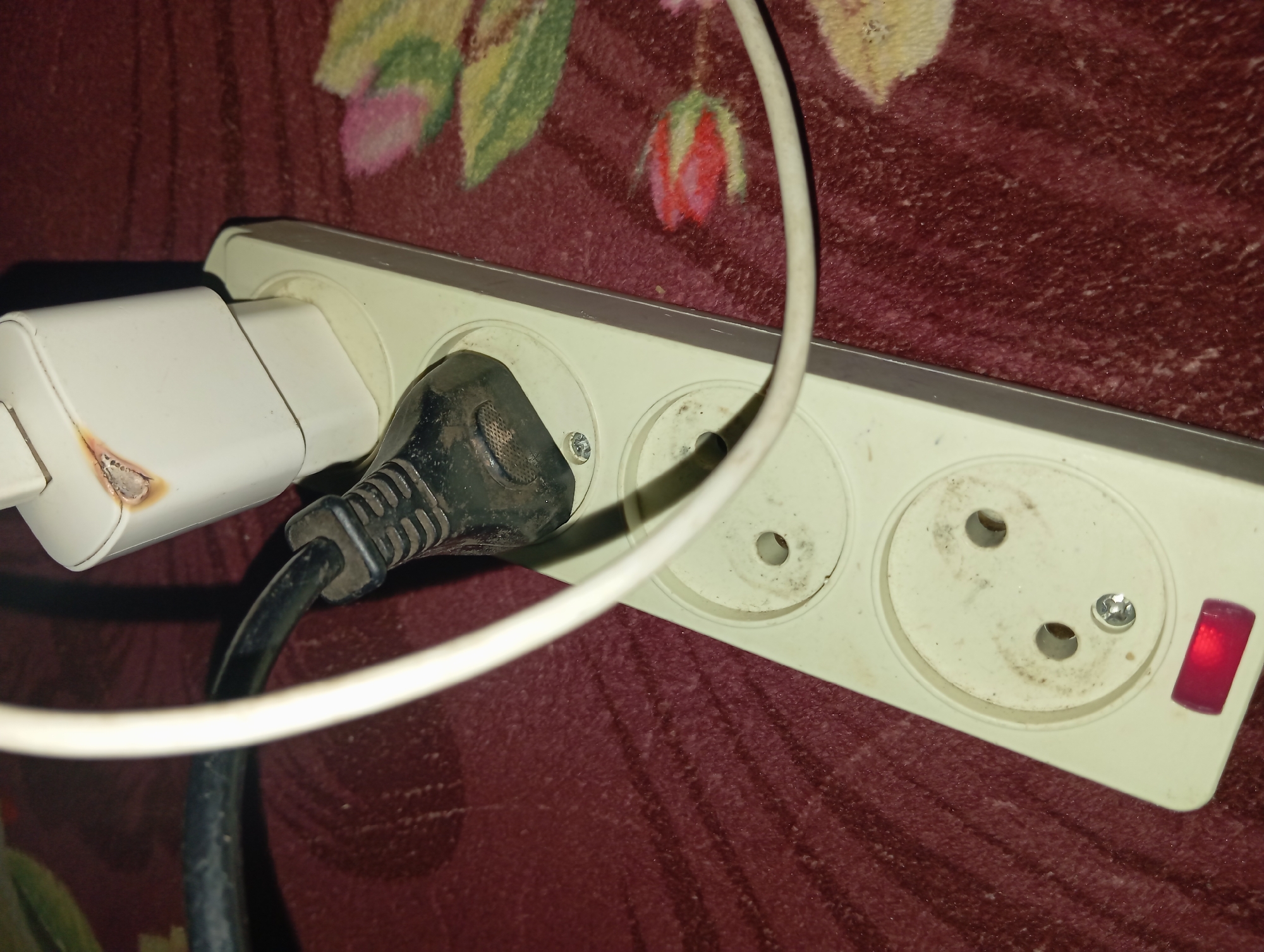 In Rejosari, Indonesia, a white electrical power strip is plugged into an outlet. A black electrical plug is inserted into one of the sockets of the power strip, and a white cable is also plugged into a socket on the power strip. The white power adapter has burn marks. The power strip is on a red carpet with a floral pattern.