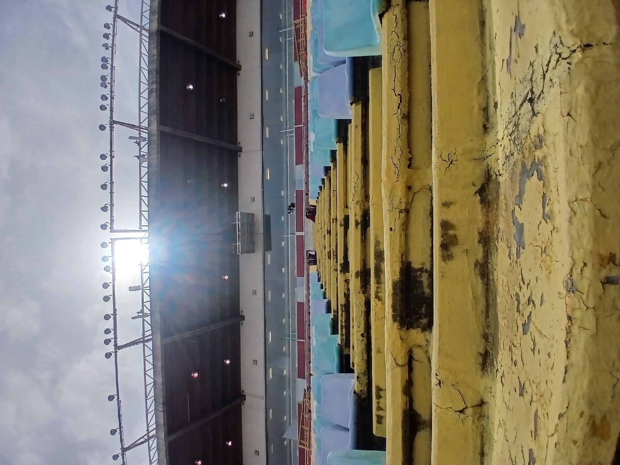 This image depicts an upward view within a stadium. The stands are characterized by yellow concrete supports with peeling paint and rows of light blue and light purple seating. On the left, a tall metal structure supporting stadium lights extends upwards into a cloudy sky. The sun is positioned behind this structure, creating a lens flare effect. In the distance, the stadium structure includes red and white walls. The location is Ciudad Guayana, Venezuela. No people are clearly visible in the image.