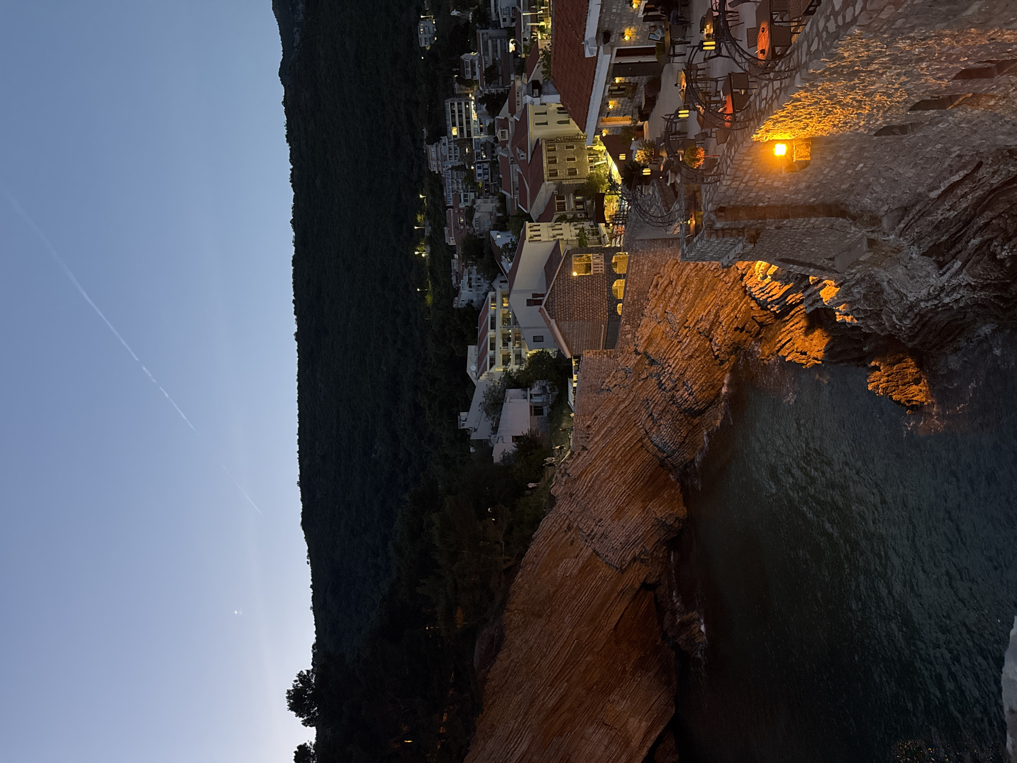 The image shows a nighttime scene in what appears to be Gornji Katun, Montenegro. A forested mountain ridge runs along the left side of the frame under a blue sky. Buildings illuminated by interior lights and streetlights cascade down the slope, suggesting a built-up area. In the foreground, rocky cliffs are illuminated by warm artificial light. The ocean is visible at the lower right. The stone architecture suggests a coastal settlement. There are no visible people or discernible actions.