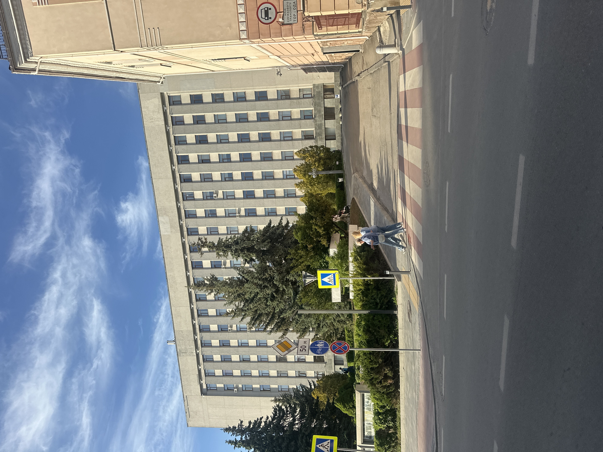 The image captures a street scene in Ternopil, Ukraine. A tall, gray building with multiple floors and many windows dominates the background. A clear blue sky with wispy clouds is visible behind the building. In the foreground, there's a road with a crosswalk featuring red and white stripes. Two individuals are seen walking across the crosswalk. Various street signs are present, including pedestrian crossing signs, a diamond-shaped warning sign, and other regulatory signs. Lush green trees and bushes line the side of the road. The overall perspective is a slightly angled view of the street, showcasing both the architectural features and the pedestrian activity.
