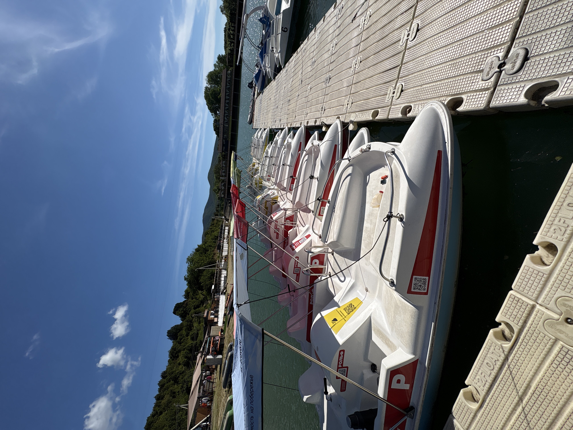 The vertically oriented image depicts a bright, sunny day at a waterfront location in Sukko, Russia, likely a lake or calm sea. A long floating pier, constructed from textured grey modular sections, dominates the mid-ground. Moored neatly along one side of the pier are numerous pedal boats, predominantly white with red accents. Each boat features a prominent red letter "P" on its side, along with a QR code and a small yellow sticker displaying the word "РОЛЬФ" (ROLF). Some pier sections also have "2F-FLOAT" embossed on them.
In the background, a lush, tree-covered shoreline rises, transitioning into distant green mountains or hills under a vast, clear blue sky adorned with scattered wispy clouds. Along the immediate shore, behind the pier, several small buildings and structures are visible, suggesting a recreational area or resort. A long white banner with partially visible Russian text, including "ность на побережье" (likely part of "opportunity on the coast"), stretches along the bank. Small groups of people can be seen enjoying the facilities on the distant shore. The scene conveys a sense of summer leisure and outdoor activity under excellent weather conditions.