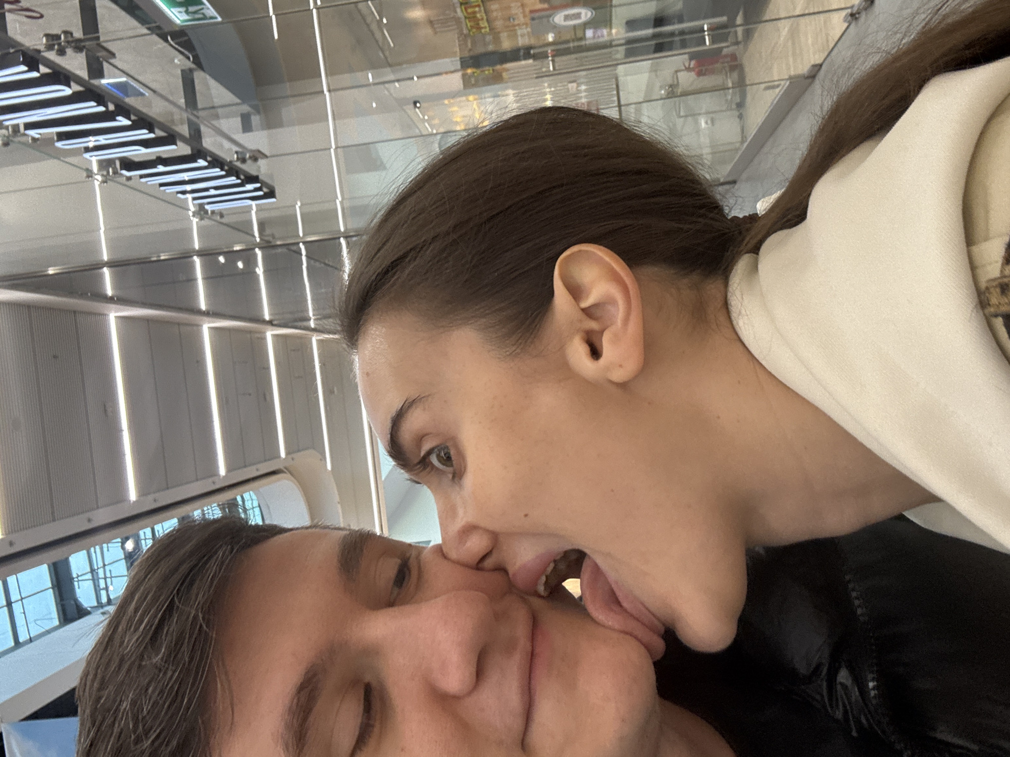 This close-up indoor photograph features two individuals, a woman and a man, engaged in a playful and affectionate interaction. The woman, positioned on the right side of the frame, is seen with her mouth open and tongue extended, playfully "licking" or nipping at the man's face, specifically his nose and cheek area. Her right eye is visible, looking towards the man. The man, on the left, has his eyes mostly closed and a slight smile, indicating he is comfortable with the playful gesture.
The woman has dark brown hair, pulled back, and is wearing a light-colored, possibly cream or white, hooded top. The man has dark hair and is wearing a dark, likely black, jacket.
The background reveals a modern indoor environment, characterized by reflective surfaces such as a glass ceiling and walls. Numerous linear lights are visible on the ceiling, along with reflections of architectural structures. Visible text in the ceiling's reflection includes inverted white neon-like letters that partially spell what appears to be "WIZARD" or "WIJAND", and a green "EXIT" sign. The bright lighting suggests it is daytime. The setting, with its contemporary design and reflective elements, is consistent with a public space like a shopping mall or airport in a city such as Warsaw, Poland. The overall mood conveyed is one of lighthearted intimacy and humor.