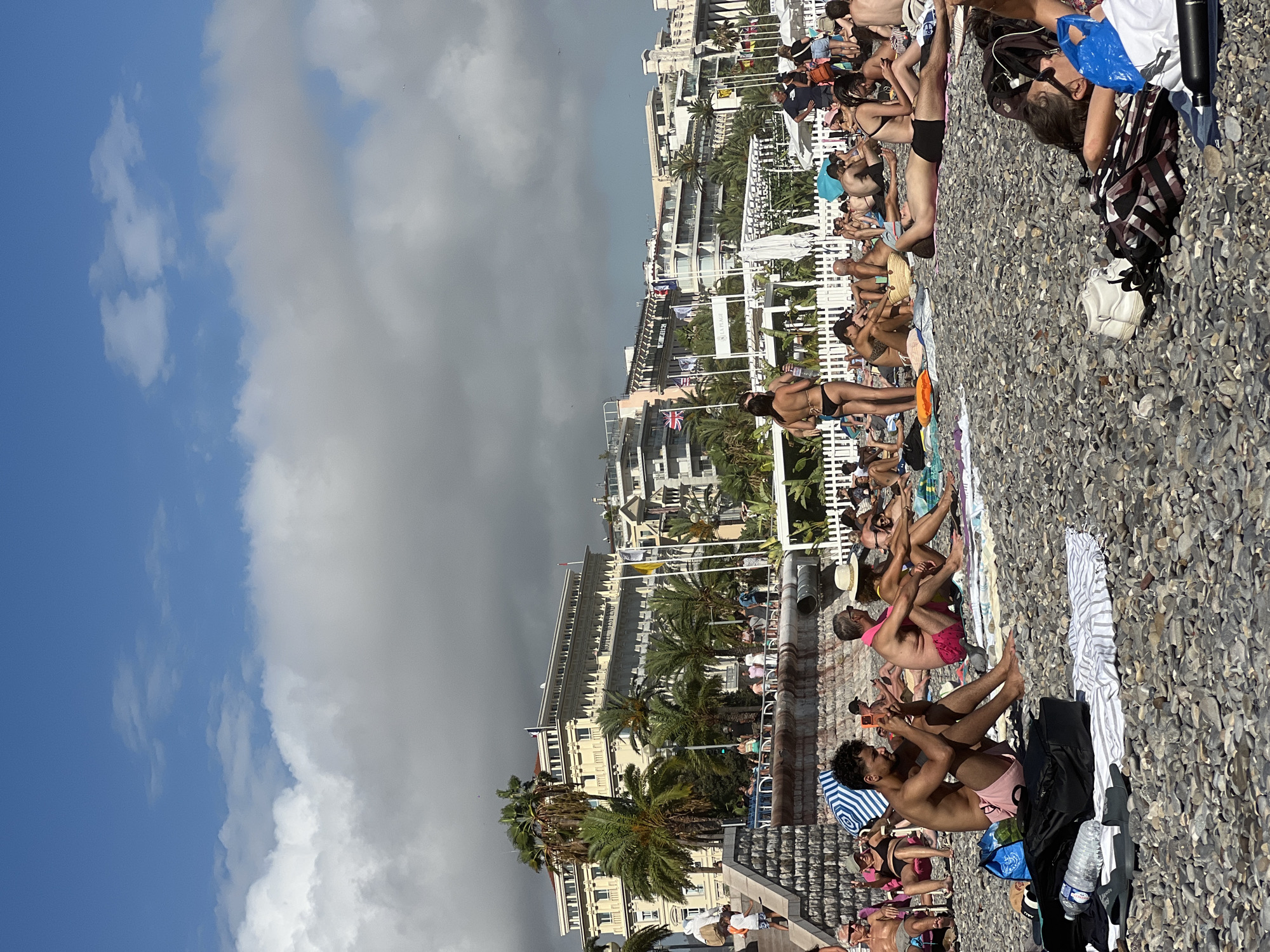 The image depicts a crowded pebble beach in Nice, France, with numerous people sunbathing and relaxing. In the background, grand hotels and palm trees line the promenade. Several individuals are lying on towels, some are sitting up, and one man is taking a photo with his phone. Beach bags, a bottle of water, and shoes are scattered on the pebbles. The sky is partly cloudy with bright sunlight.