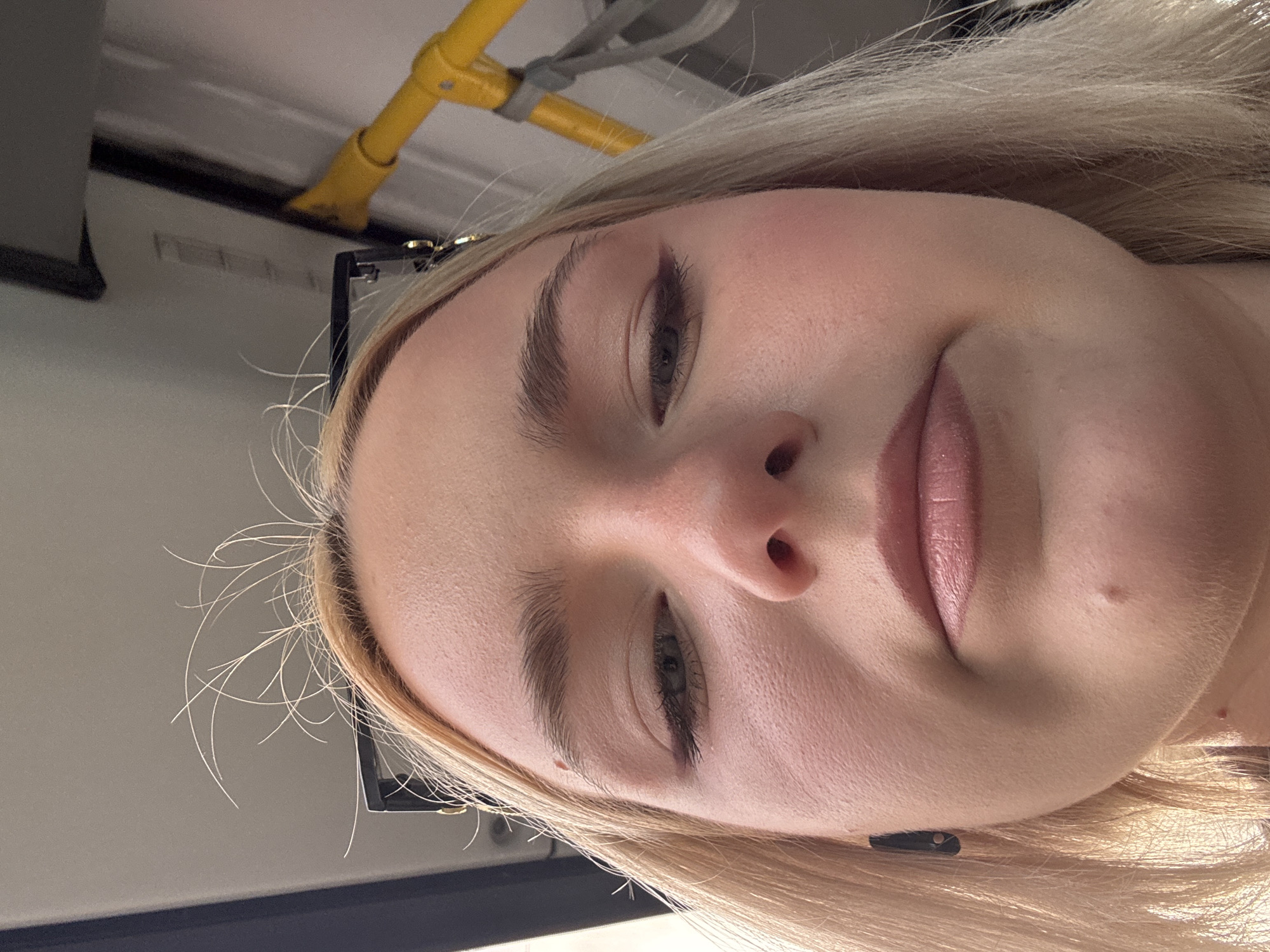 The image shows a close-up, low-angle shot of a young blonde woman's face, taken from what appears to be inside a vehicle, likely public transportation. Her eyes are partially closed, and her expression is neutral. She is wearing makeup, including eyeliner and lip gloss. A small black earbud is visible in her left ear. Stray blonde hairs are sticking up from her head. In the background, parts of the vehicle's interior are visible, including a yellow handrail and a grey ceiling panel. The overall lighting suggests it is daytime. The location is identified as Sinyavino, Russia, however, there are no visual elements in the image that specifically confirm this location.