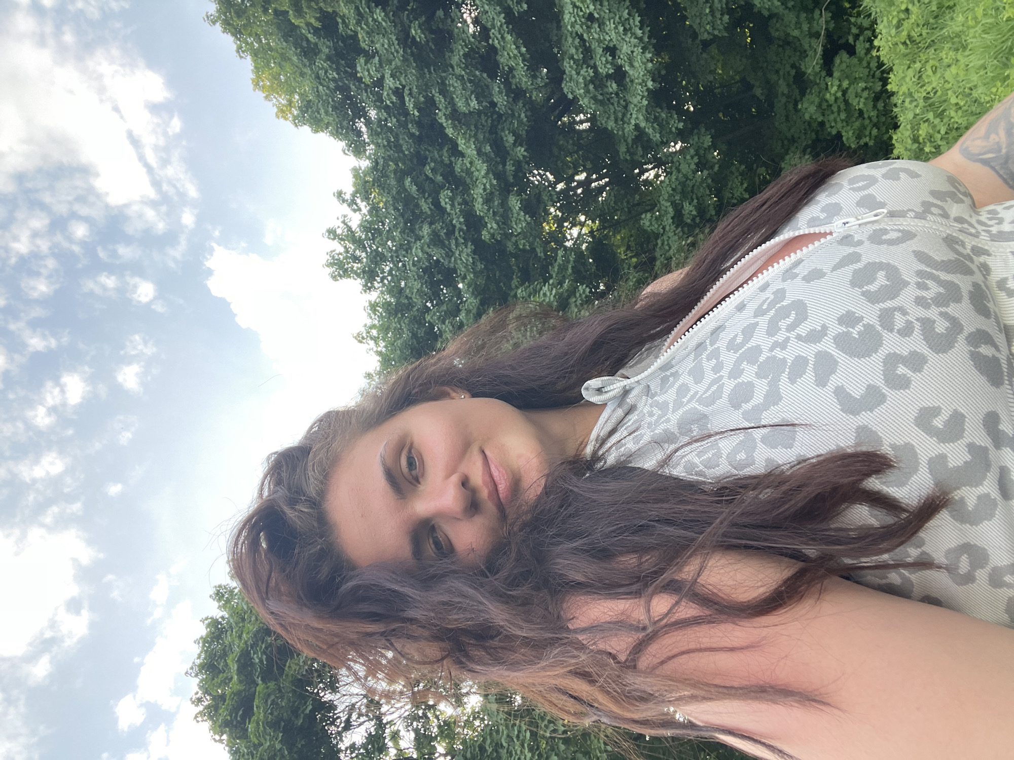 A woman with long brown hair looks directly at the camera. She wears a gray and white animal print zippered top. The background includes a sky with clouds and green trees. The photo's location cannot be determined from the image and context provided.