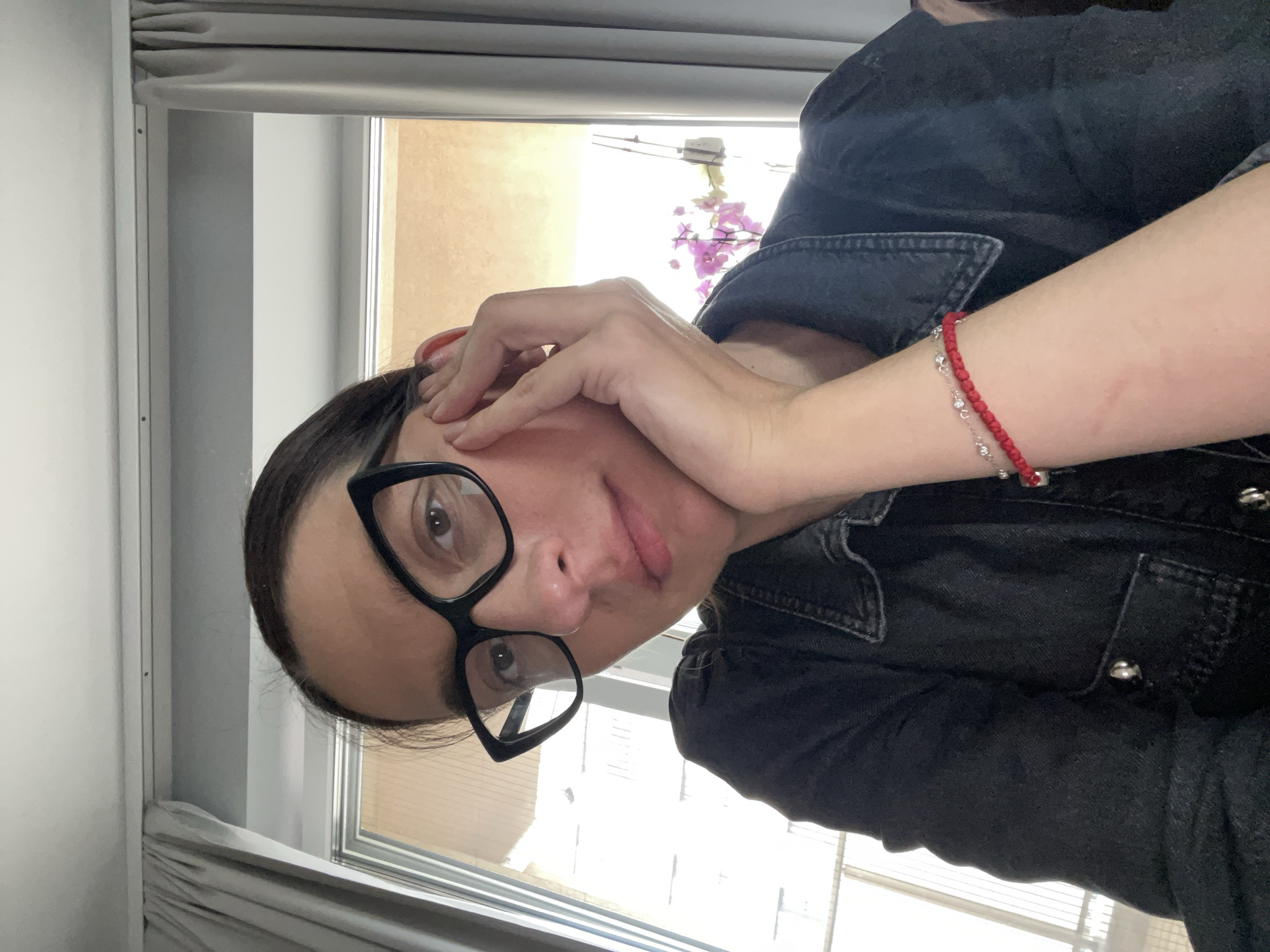 A woman with dark hair pulled back, wearing black-rimmed glasses and a dark denim jacket, is posing for a selfie. She is holding her hand up to her face, with a red and silver bracelet visible on her wrist. Behind her is a window with closed blinds and a view of what appears to be a light-colored building and some flowering plants. The location cannot be determined to be in Sofia, Bulgaria based on the image provided.