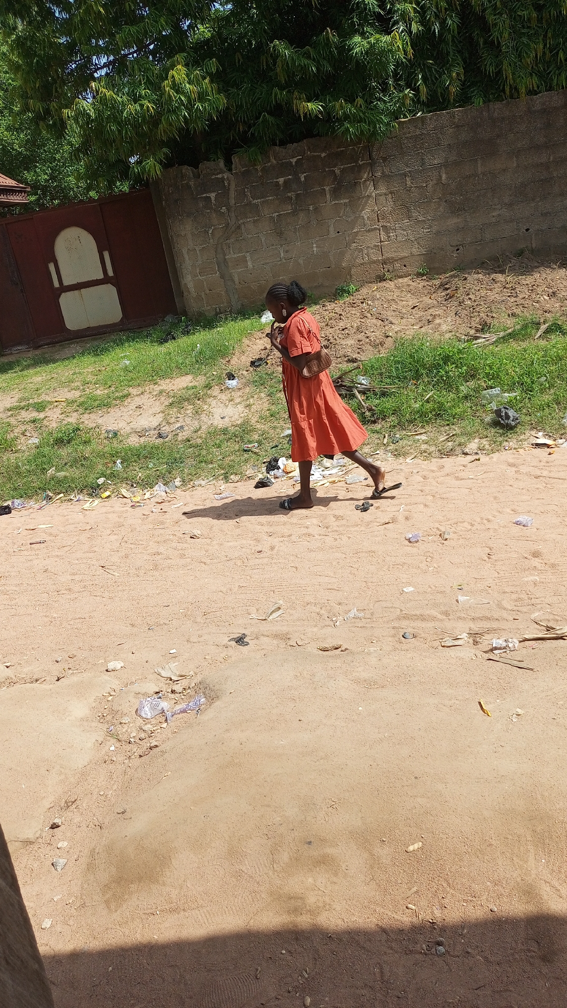 In Jalingo, Nigeria, a young girl in an orange dress walks along a dirt path. She is wearing dark sandals and carrying a small brown purse over her shoulder. Her hair is pulled back. She appears to be looking down at something in her hand as she walks. The ground is sandy and littered with trash, including plastic bags and wrappers. To the left, there is a tall red gate with a decorative design, and behind it, green trees. A rough concrete wall runs along the background, partially covered by the trees. The overall environment suggests an outdoor area, possibly a roadside or an undeveloped part of the city.