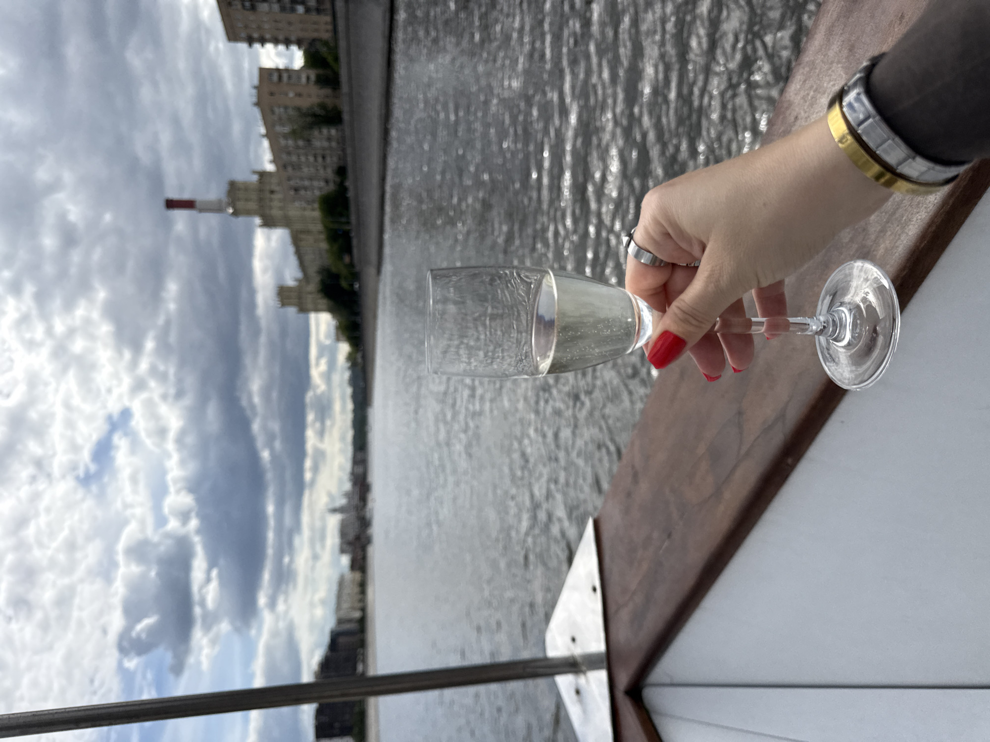 A person holds a glass of champagne in their right hand on a boat. The person has red nail polish, a ring on the ring finger and two bracelets on the wrist. The drink is clear and bubbly. Behind the person, there is the Moscow River. The cityscape on the left is the embankment of the Novodevichy Convent in Moscow, Russia. The sky is overcast.