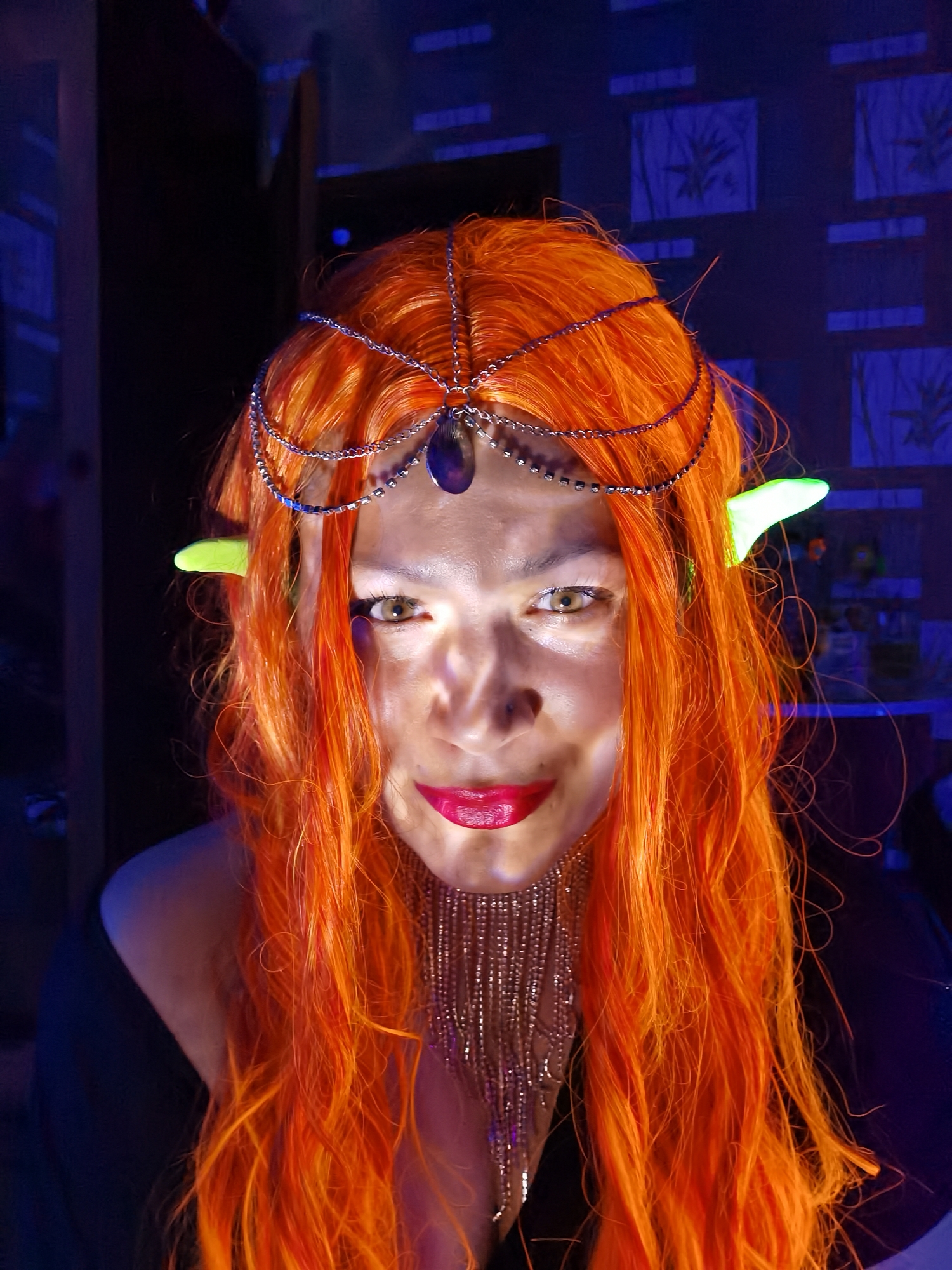 The image shows a woman with orange hair and pointed, possibly prosthetic, green ears. She is wearing a silver headpiece with a dark gemstone and a beaded necklace. Her lips are painted red. The background is dark, but shows wallpaper with a subtle floral pattern. The lighting is unusual, with a strong light source shining directly onto the woman's face. No evidence indicates that the photo was taken in the city of Chaikovsky, Russia.