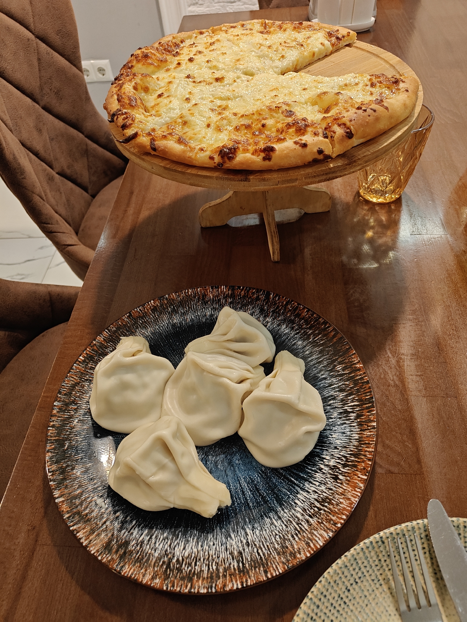 A culinary scene unfolds indoors in Batumi, Georgia. A table dominates the frame, hosting a feast of Georgian cuisine. A large, round Adjarian khachapuri, a traditional cheese bread, rests on a wooden stand with a slice already consumed. Next to it stands a faceted glass. A plate of khinkali, Georgian dumplings, lies in the foreground, their twisted tops facing up. Cutlery and a patterned plate are visible in the lower right corner. A brown chair occupies the left side of the shot, partially visible, along with a glimpse of tiled flooring. The table is made of wood. The lighting is diffused, creating a warm ambiance.