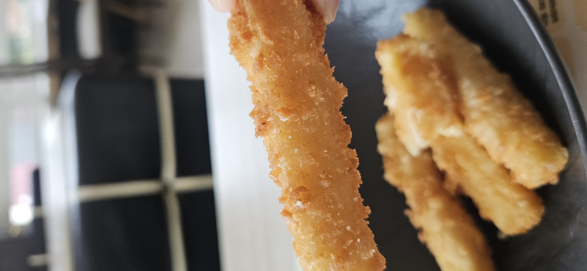A close-up, vertical shot shows a person holding a fried food item, possibly a mozzarella stick or a piece of fried chicken, in the foreground. The food item is golden brown and coated in a crispy breading with visible salt crystals. The background is blurred, but suggests an indoor setting with dark seating and a light-colored surface. The food item appears to be part of a plate of similar fried items in the lower portion of the frame. No specific location in Vitsebsk, Belarus can be identified from this image.