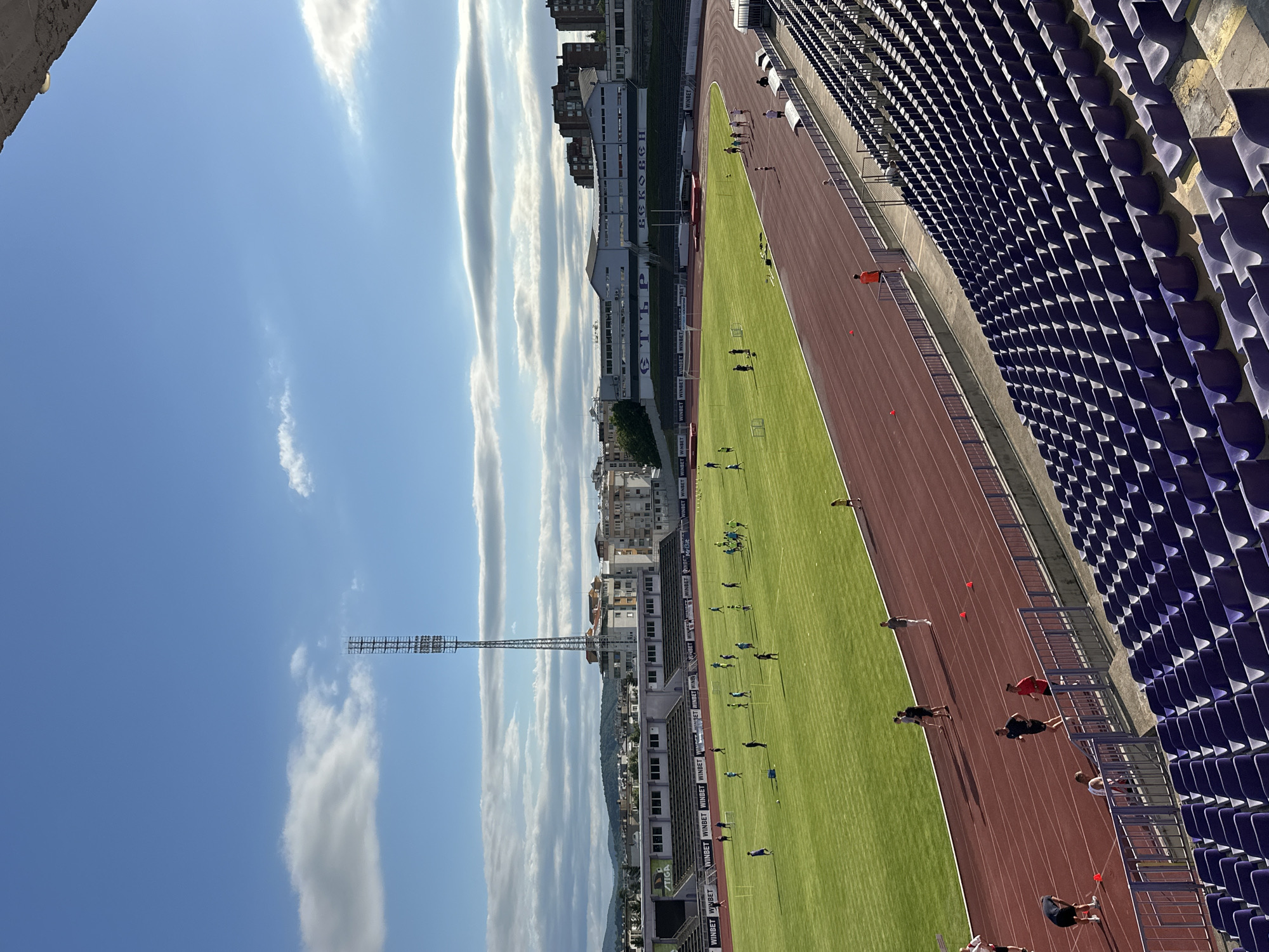 The image depicts a view of the Ivaylo Stadium in Veliko Tarnovo, Bulgaria, on a sunny day with scattered clouds. The stadium features a lush green football pitch and a red athletic track. Several individuals, presumably athletes, are engaged in training activities on both the field and the track. Some are running, while others are standing or walking. Spectator stands, mostly empty and upholstered in purple, are visible in the foreground. In the background, buildings and a tall transmission tower are set against a backdrop of rolling hills and a blue sky. The overall scene captures a moment of athletic practice at the stadium.