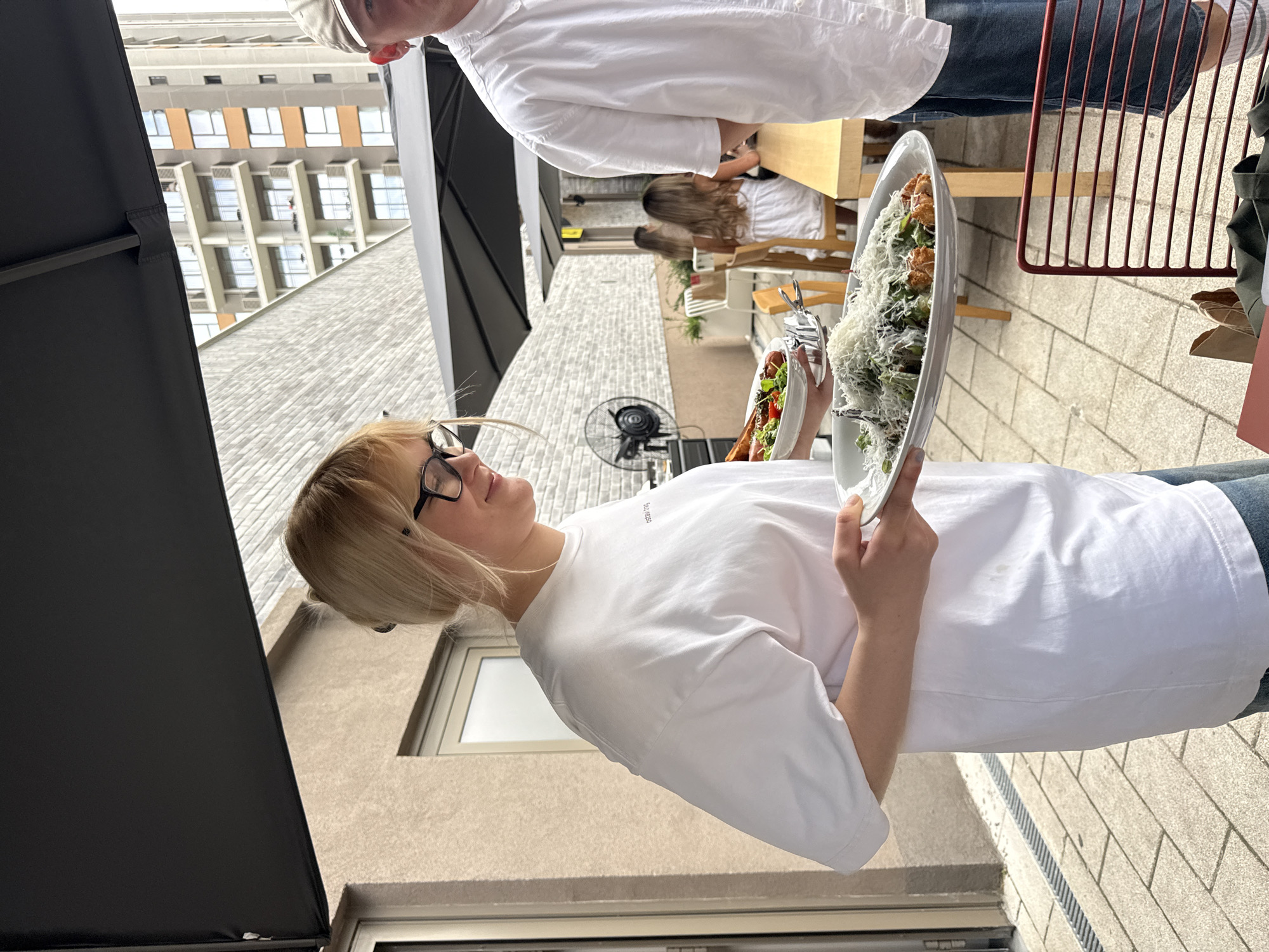 In Belgrade, Serbia, a young woman with blonde hair and glasses, wearing a white t-shirt and jeans, holds two plates of food. The plate in her left hand contains a salad with tomatoes and greens, along with some sausages. The plate in her right hand features what appears to be fried food topped with grated cheese, possibly pasta or vegetables. She is standing outdoors on a paved patio, likely part of a restaurant or cafe, as suggested by the tables and chairs visible in the background, as well as a large black umbrella providing shade. A man in a white shirt and a cap is partially visible to her right. The background shows a modern brick building and another building with multiple balconies, indicating an urban setting. The overall scene depicts a casual dining experience in an outdoor urban environment.