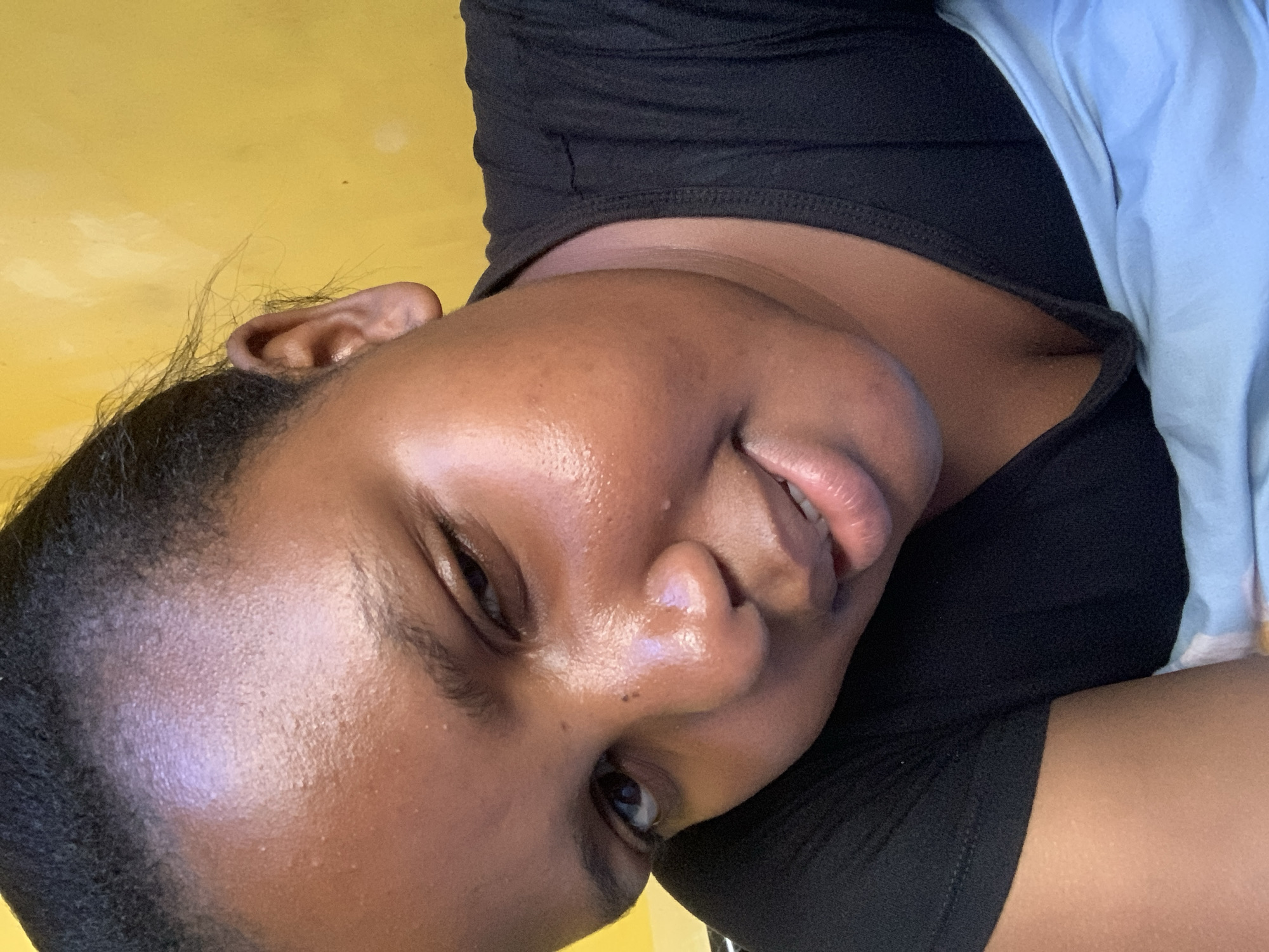A close-up photo features a dark-skinned woman with glossy skin. She is smiling slightly with her lips parted. Her dark hair is neatly pulled back. She is wearing a black top and a light blue or white garment partially visible on the side. The background is a plain yellow surface. No location information is discernible in the image.