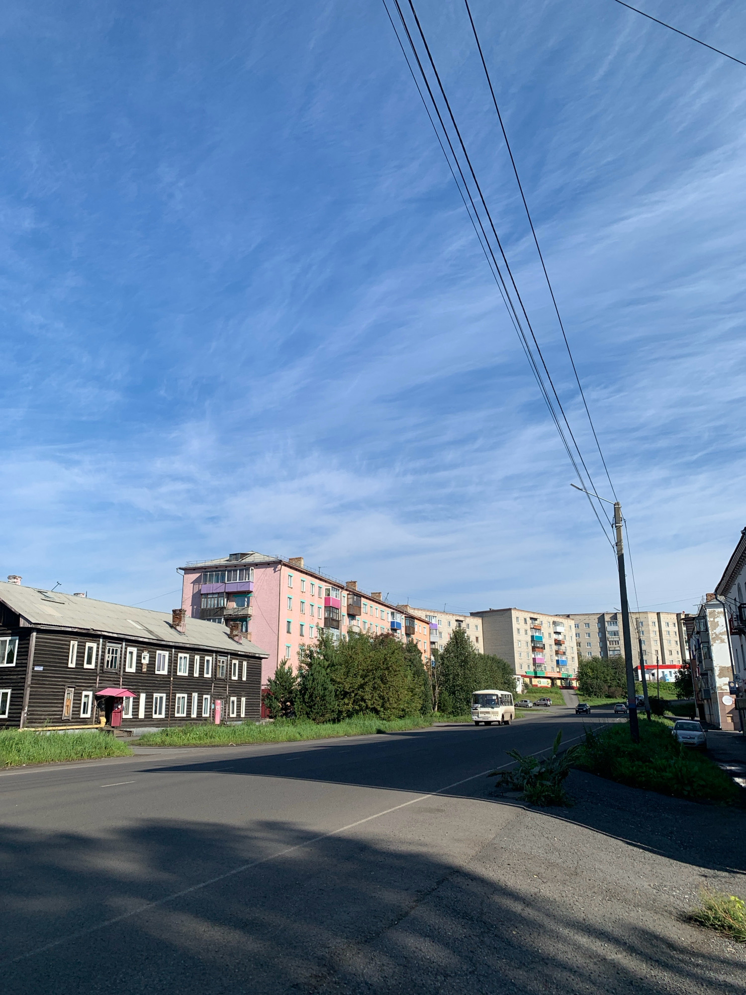 The image shows a street scene in Prokopyevsk, Russia, on a bright, clear day with a sky full of wispy clouds. On the left side of the street, a long, dark wooden building with many windows and a metal roof stands. Adjacent to it is a tall, pink apartment building with colorful balconies. Further down the street, a row of light-colored apartment buildings with balconies and windows can be seen. A white bus is driving down the street, and a few cars are parked or driving in the distance. Power lines stretch across the sky, supported by a telephone pole. The street is paved, and there are areas of grass and trees lining the sides. The overall impression is of a typical residential street in a Russian city.