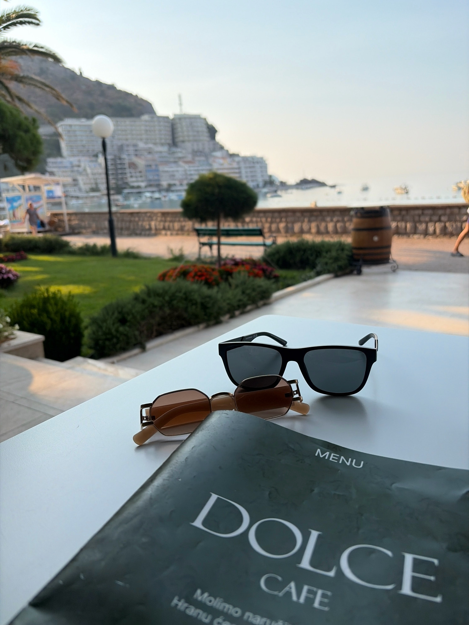 In Bečići, Montenegro, on a white table in front of a cafe, two pairs of sunglasses and a menu are placed. The sunglasses are positioned in the foreground, with a larger black pair behind a smaller gold-rimmed pair. The menu, predominantly dark green with white lettering, is partially visible, showing the words "MENU," "DOLCE," and "CAFE." In the background, a lush green lawn with flowerbeds separates the table from a stone walkway. Beyond the walkway, a large wooden barrel stands near a stone wall, with a glimpse of a resort town built into a hillside overlooking the sea. The sky is clear and bright, indicating it is daytime.