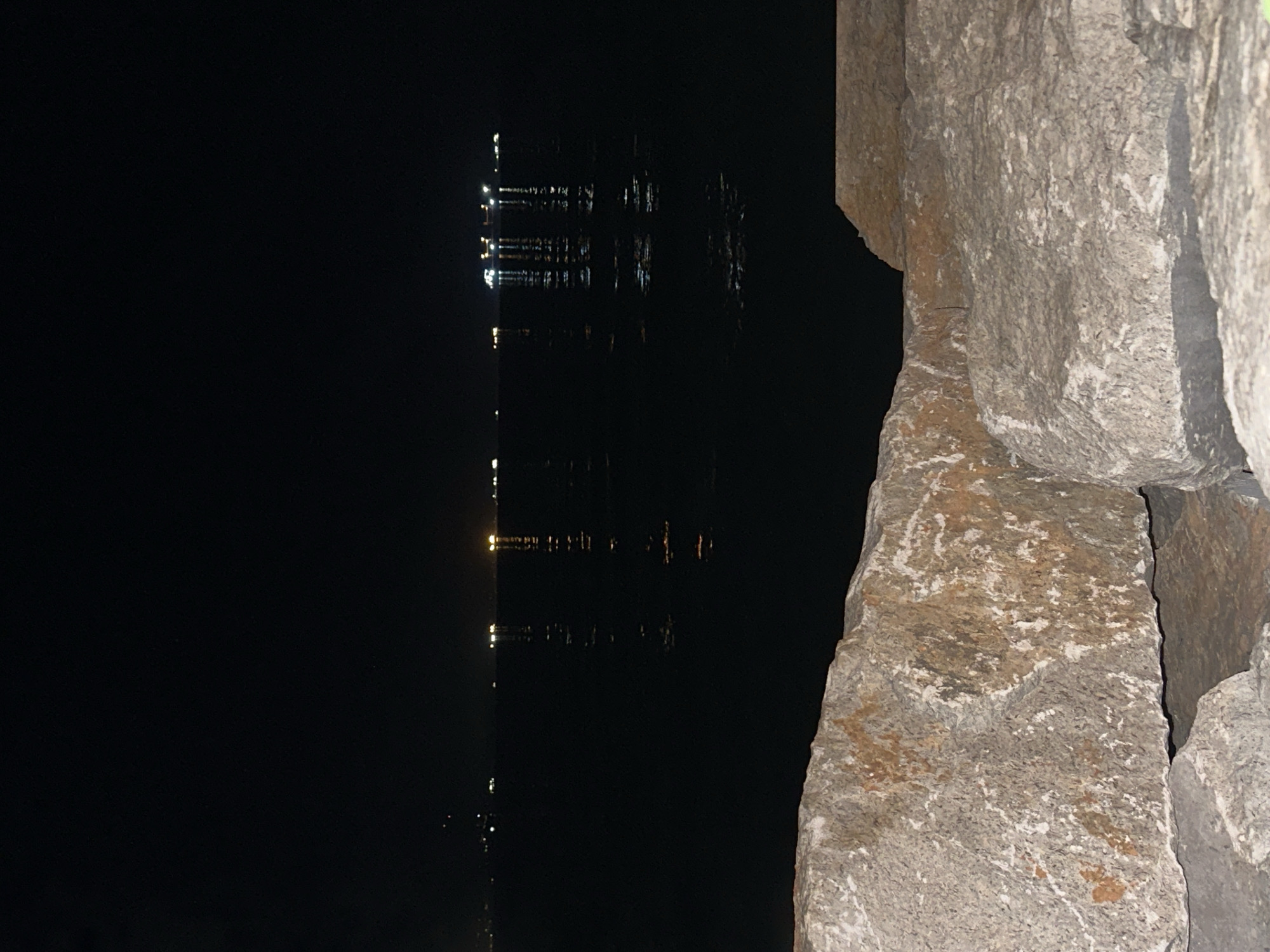 The image shows a nighttime view of the sea with lights from ships and possibly a distant shoreline visible on the horizon. In the foreground, large rocks are in sharp focus, textured and varied in color, with patches of white and rust-colored mineral deposits. The water is dark and still, reflecting the distant lights as vertical streaks. The sky is completely black, indicating nighttime. The scene is likely a coastal area, possibly a harbor or bay. The prompt specifies that the location is the city of Trieste, Italy.