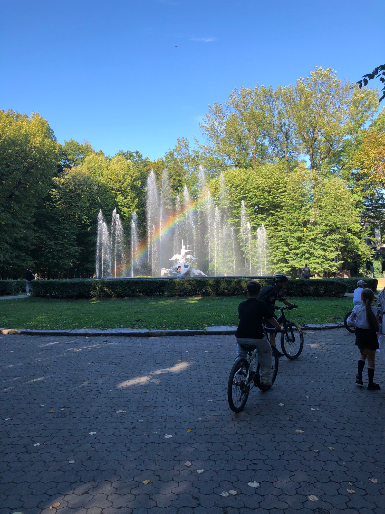 This is a photograph of the Fountain of Neptune in Lviv, Ukraine, located in the city center. The fountain is in the background, with water jets shooting upwards, creating a rainbow. There are people in the foreground riding bicycles and walking. The ground is paved with interlocking stones and scattered leaves. The trees surrounding the fountain are lush and green, indicating it is likely a summer or early autumn day. The sky is a clear blue, with a single airplane visible in the upper portion.