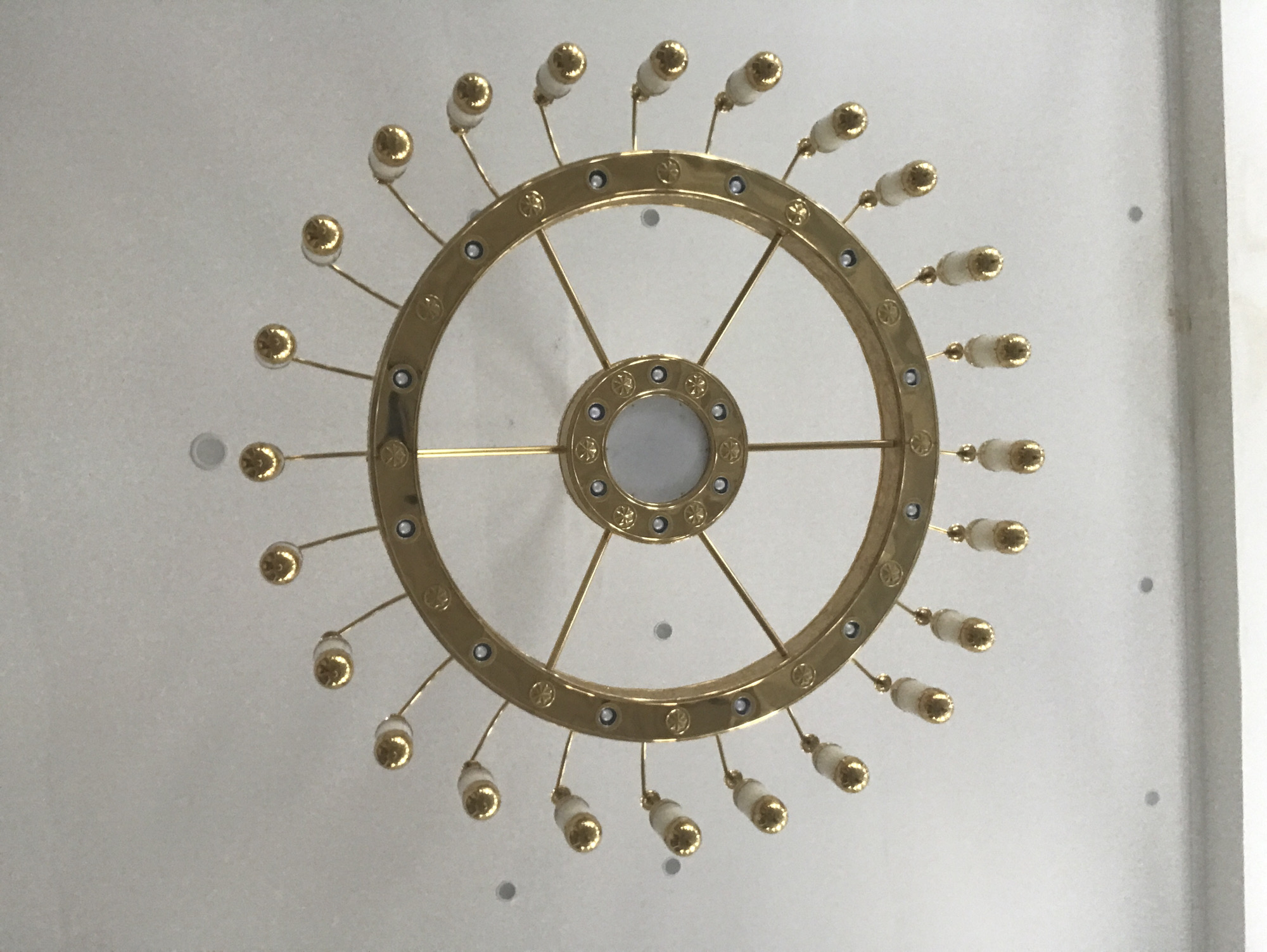 The image shows a chandelier attached to a white ceiling. The chandelier is circular, made of gold-colored metal, with a central, round, decorative element and radiating spokes connecting to the outer ring. The outer ring holds numerous small, bulb-shaped fixtures, possibly for lights. The background ceiling is plain white with some slight variations in texture and shade. The photograph is taken from directly below, looking upwards at the chandelier. The location, Meunasahaluedrienlhoksukon Barat, Indonesia, is not apparent from the image itself.