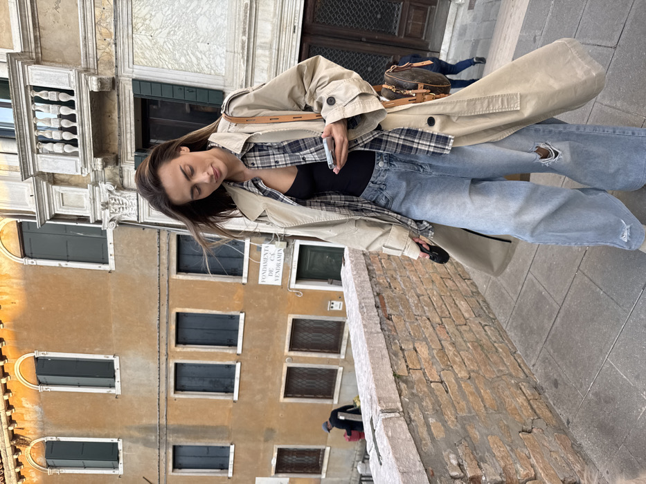 A woman is depicted walking down a stone staircase or ramp in Venice, Italy, specifically identifiable by the "FONDAMENTA DE CA' VENDRAMIN" street sign on a building in the background. The woman, appearing to be in her late 20s or early 30s with long brown hair, is looking downwards at a silver-colored smartphone held in her left hand. She wears a beige trench coat unbuttoned over a blue and white plaid shirt, which is also unbuttoned, revealing a black tank top or bodysuit. Her lower attire consists of light blue, distressed wide-leg jeans with visible rips at the knees. A brown shoulder bag with a distinctive monogram pattern hangs from her right shoulder. In her right hand, she holds a small black object, possibly a phone or sunglasses.
To the left, a weathered multi-story building with an ochre-orange stucco facade stands prominently, featuring several windows with dark shutters. Some upper windows are arched, while lower ones are rectangular. Adjacent to it on the right is a section of a lighter-colored, ornate building with a decorative stone balcony supported by corbels and featuring balusters.
In the far background to the left, a man wearing a dark hat and jacket is partially visible behind a brick wall, carrying a red container. To the far right, partially obscured, the lower legs of another individual are visible on a descending path. The foreground includes a brick wall with a stone coping, suggesting a bridge or elevated walkway. The ground surface is composed of paved stone. Natural light illuminates the scene, highlighting the architectural details and the warm tones of the buildings.