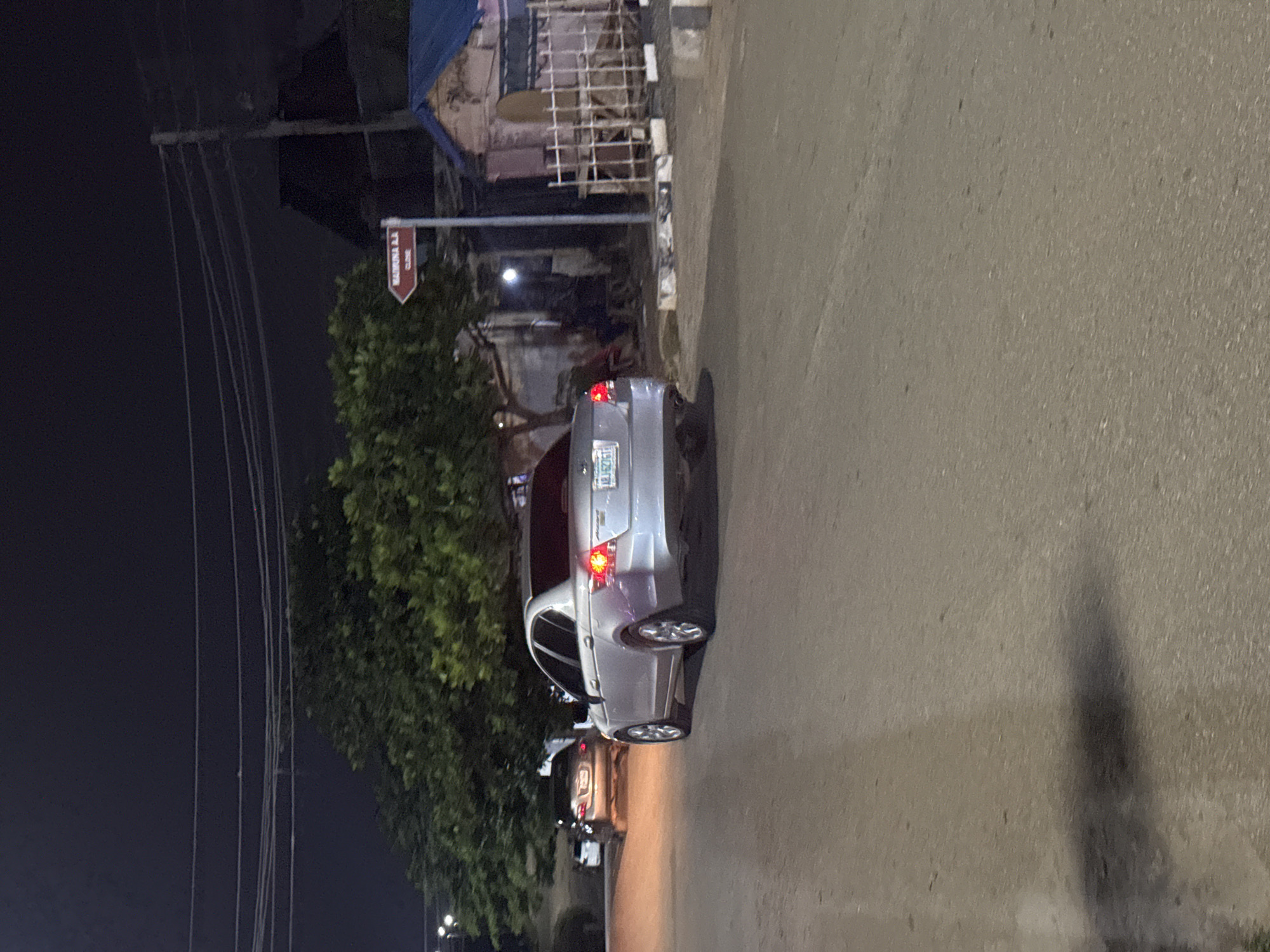 The image depicts a nighttime street scene in Abuja, Nigeria. A silver sedan, possibly a Honda Accord, is prominently featured in the foreground, driving away from the camera. The car's taillights are illuminated, and its license plate is visible, though partially obscured. In the background, another car is seen driving towards the camera, its headlights casting an orange glow on the road.
To the right of the street, a weathered building with a blue corrugated metal roof stands adjacent to a metal fence. A red directional sign is also visible, pointing to "MADRINA CLO." The environment appears to be a residential or commercial area, with trees and overhead power lines suggesting a typical urban setting in Abuja. The overall atmosphere is dimly lit, characteristic of a night scene.