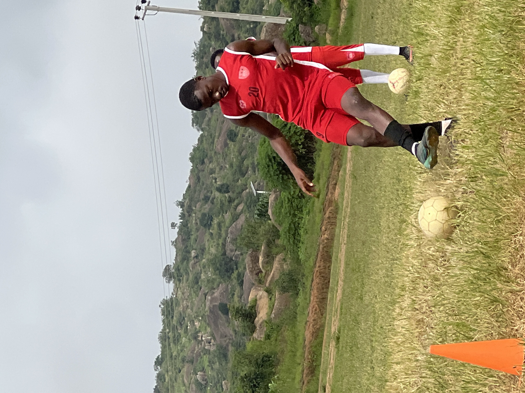 In Jos, Nigeria, a young man in a red soccer uniform with white trim, numbered "20," practices on a grassy field. He is actively kicking a soccer ball, his right leg extended. Another player in a similar uniform stands behind him. The field is covered in dry, yellowing grass and is marked with an orange cone in the foreground. The background features a hilly landscape with scattered rocks and green vegetation, under an overcast sky. A power pole with wires is visible to the right of the frame.