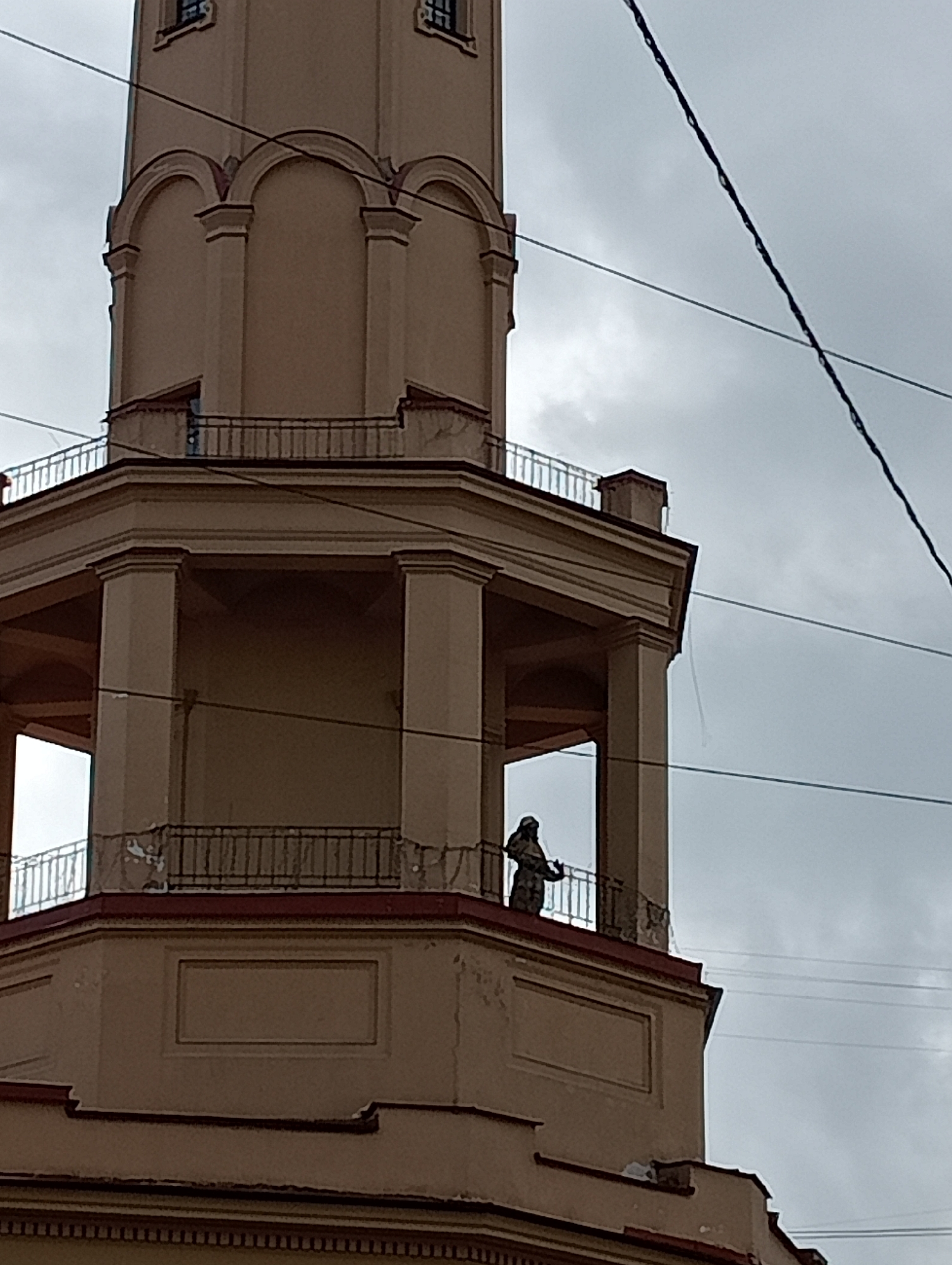 The image captures a close-up view of the upper section of a light brown, multi-tiered tower-like structure, likely a part of a historic building in Saint Petersburg, Russia. The tower features classical architectural elements, including arched windows and columns. A prominent feature is a balustraded open-air walkway or balcony encircling the second visible tier from the top. On this balcony, a lone figure, appearing to be a statue or a person dressed in dark clothing, stands looking outwards. The structure is partially obscured by a network of overhead power lines that crisscross against a heavily overcast, pale gray sky. The building itself shows signs of age, with some areas of chipped paint and weathering visible on the façade. The perspective is from below, looking upwards.