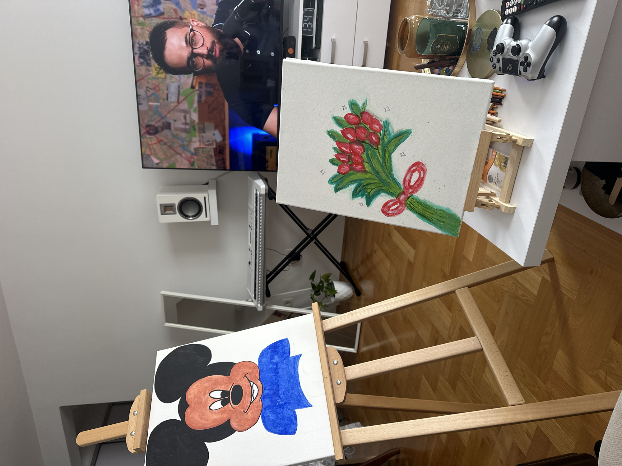 The image depicts an indoor scene featuring a living space with a television mounted on a white wall. The TV screen displays a man with glasses, possibly from a broadcast or recording. Below the TV is a white speaker. Next to the speaker is a white storage unit. An easel holds a canvas painting of a bouquet of red berries tied with a red ribbon. On a nearby white table are items including mugs, a tray with glasses, a video game controller, and art supplies. Another easel holds a canvas painting of Mickey Mouse. The floor is wooden and reflects light. The location within the city of Budva, Montenegro is not discernible from the visual information provided.