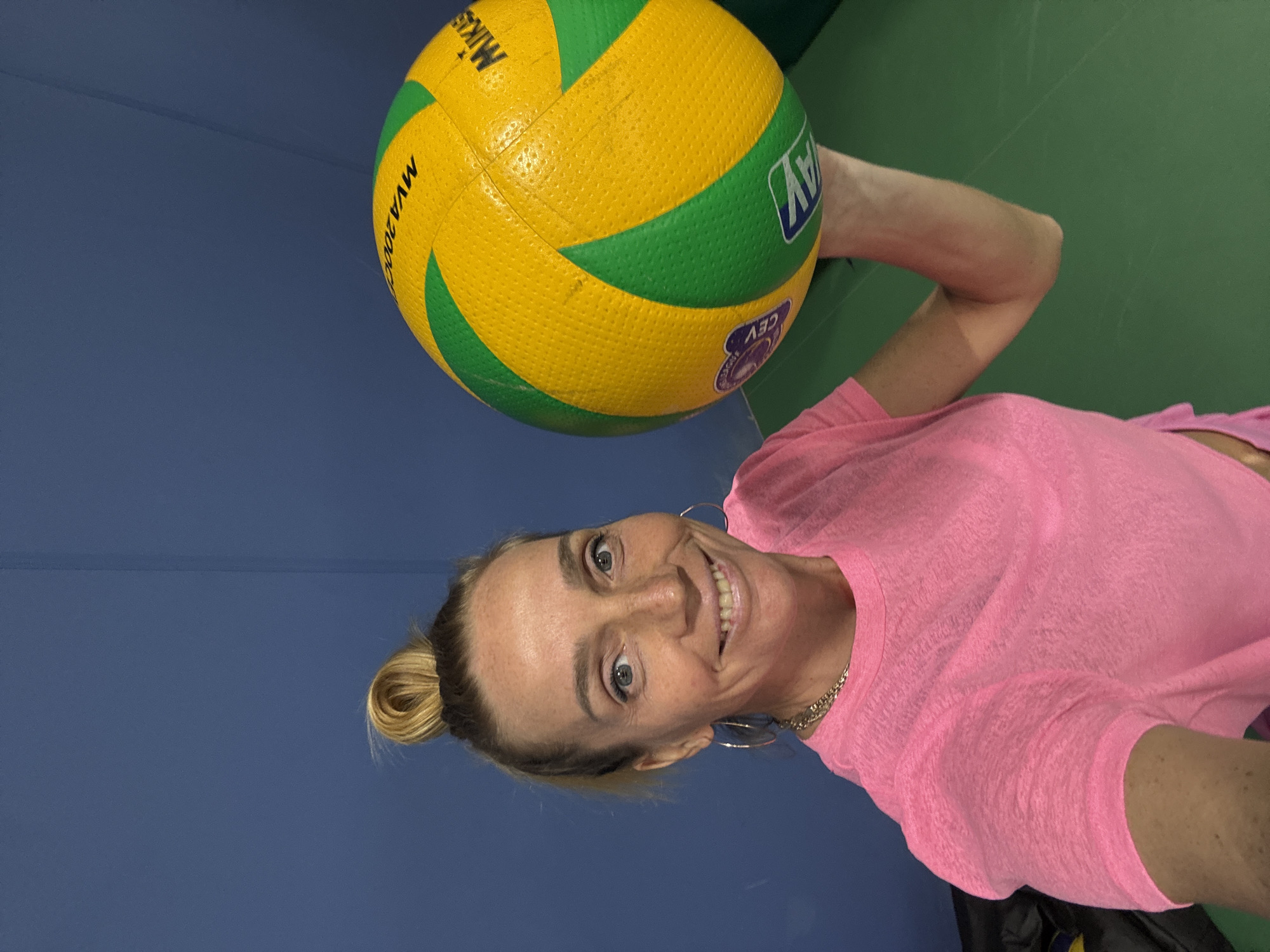 A woman in a pink shirt holds a yellow and green volleyball. The location appears to be indoors with blue walls and green flooring. There is no information available that would place this image in Saint Petersburg, Russia.