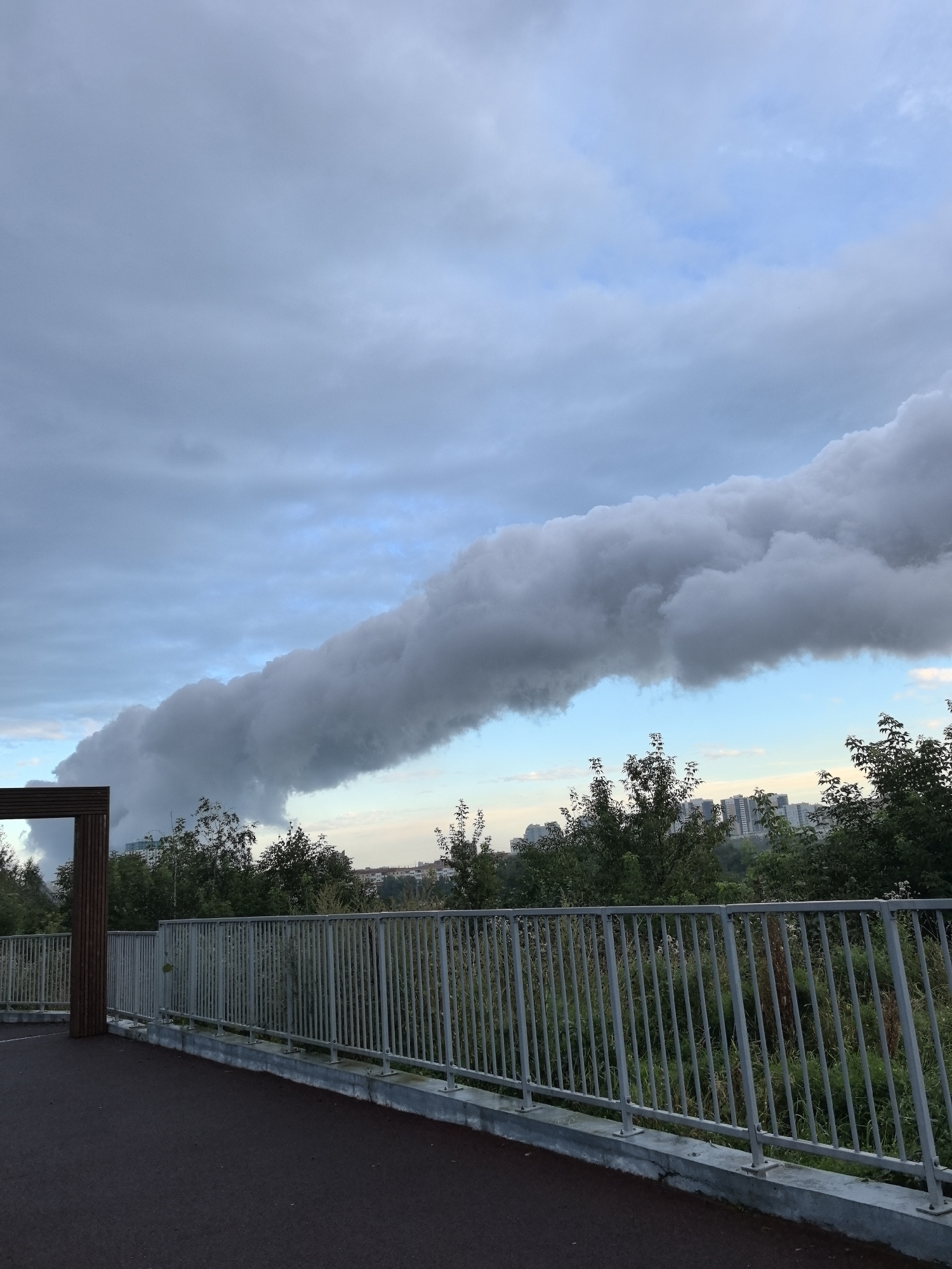 The image shows a view from a pedestrian bridge in Perm, Russia. A massive, dark cloud formation stretches across the sky, obscuring a significant portion of the horizon. Below the cloud, a line of green trees separates the foreground from a cityscape in the distance, characterized by apartment buildings. A silver metal railing runs along the side of the bridge, and a dark paved surface is visible in the foreground. A wooden structure, possibly part of an archway or seating area, is located on the left side of the bridge. The sky is a mix of grey clouds and patches of blue.