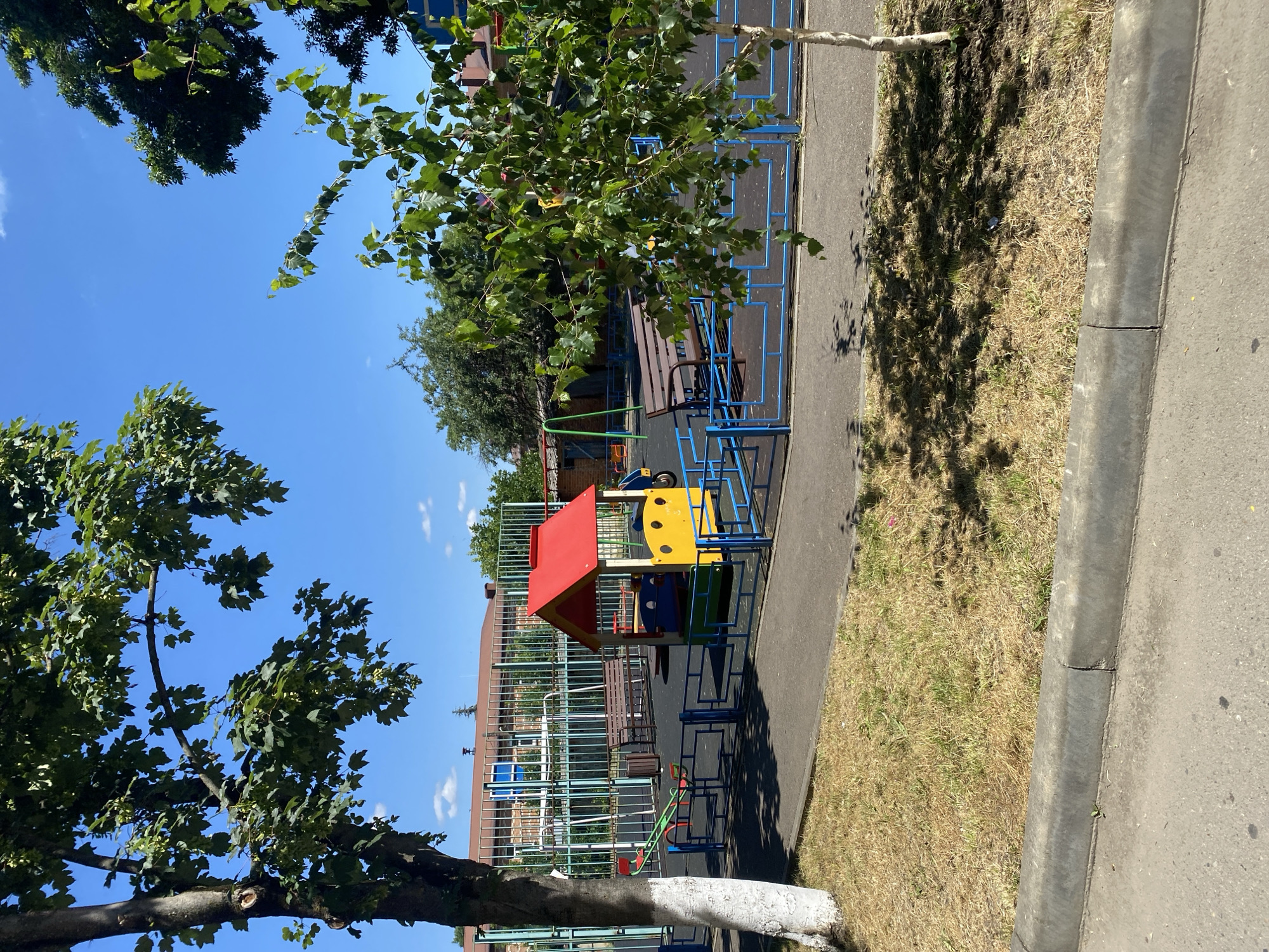 A brightly colored playground with a small red-roofed playhouse, a seesaw, and a swing set is visible under a clear blue sky on a sunny day. The playground is enclosed by a blue metal fence, and mature trees with green leaves surround the area. In the background, a building with a brick facade is partially visible behind a tall metal fence. The ground is covered with black rubber safety surfacing in the playground area, while the surrounding areas have dry, yellowed grass and asphalt pathways. A concrete curb runs along the edge of the asphalt in the foreground. The scene appears to be located in a residential or public park area.