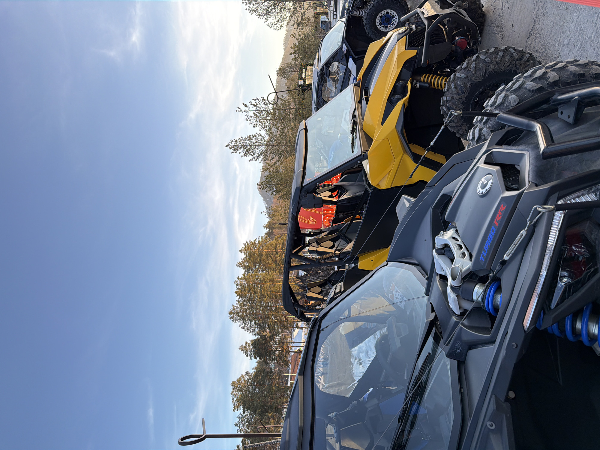 The image shows a row of three all-terrain vehicles (ATVs), presumably in the city of Озёрное, Russia. The ATVs are parked close together, with the front of each vehicle facing the viewer. The first ATV is dark gray, with "TURBO RR" branding visible. The second ATV is yellow, and the third appears to be black. In the background, there are trees, a building, a fence, and a utility pole. The sky is blue with some clouds.