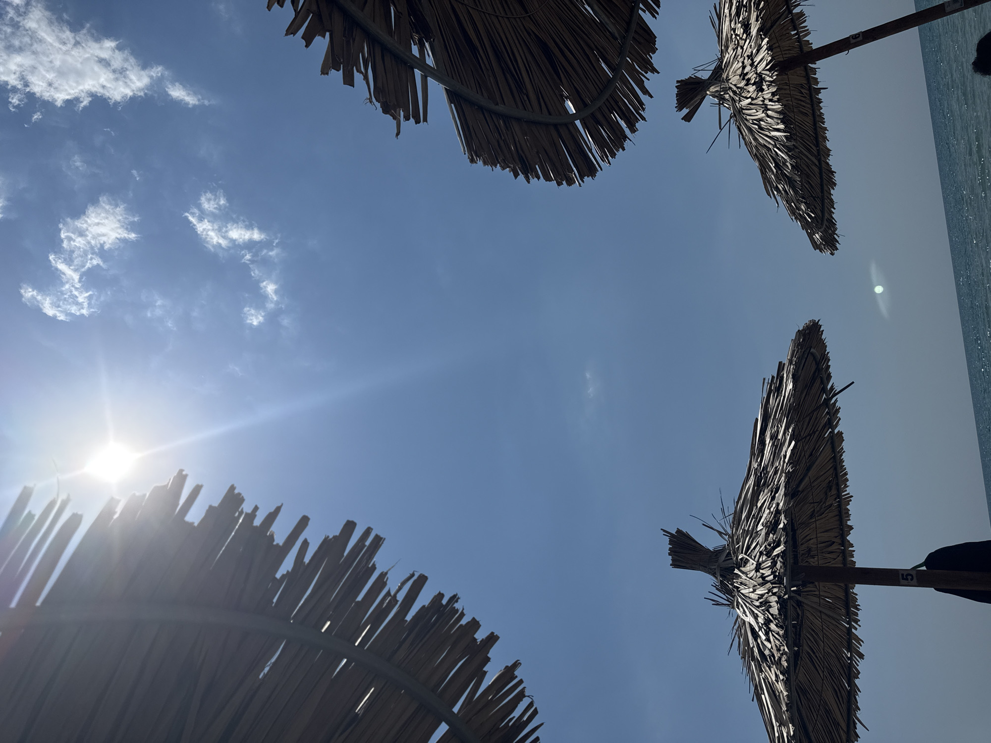 The image shows a bright, sunny sky with a few wispy clouds. The sun is high in the sky, casting strong light and creating lens flare. In the foreground, there are several thatched beach umbrellas, made of natural materials like straw or palm leaves. These umbrellas are angled towards the viewer from different perspectives, with the one on the left taking up a significant portion of the frame. Some of the umbrellas have visible poles and one has a black bag or clothing item hanging from it, with a number "5" visible on the pole. The umbrellas are arranged as if they are on a beach, with a hint of the sea and ocean waves visible in the background at the bottom of the frame. The overall impression is of a typical summer day at a beach, likely in the city of Imeros, Greece, given the context provided.