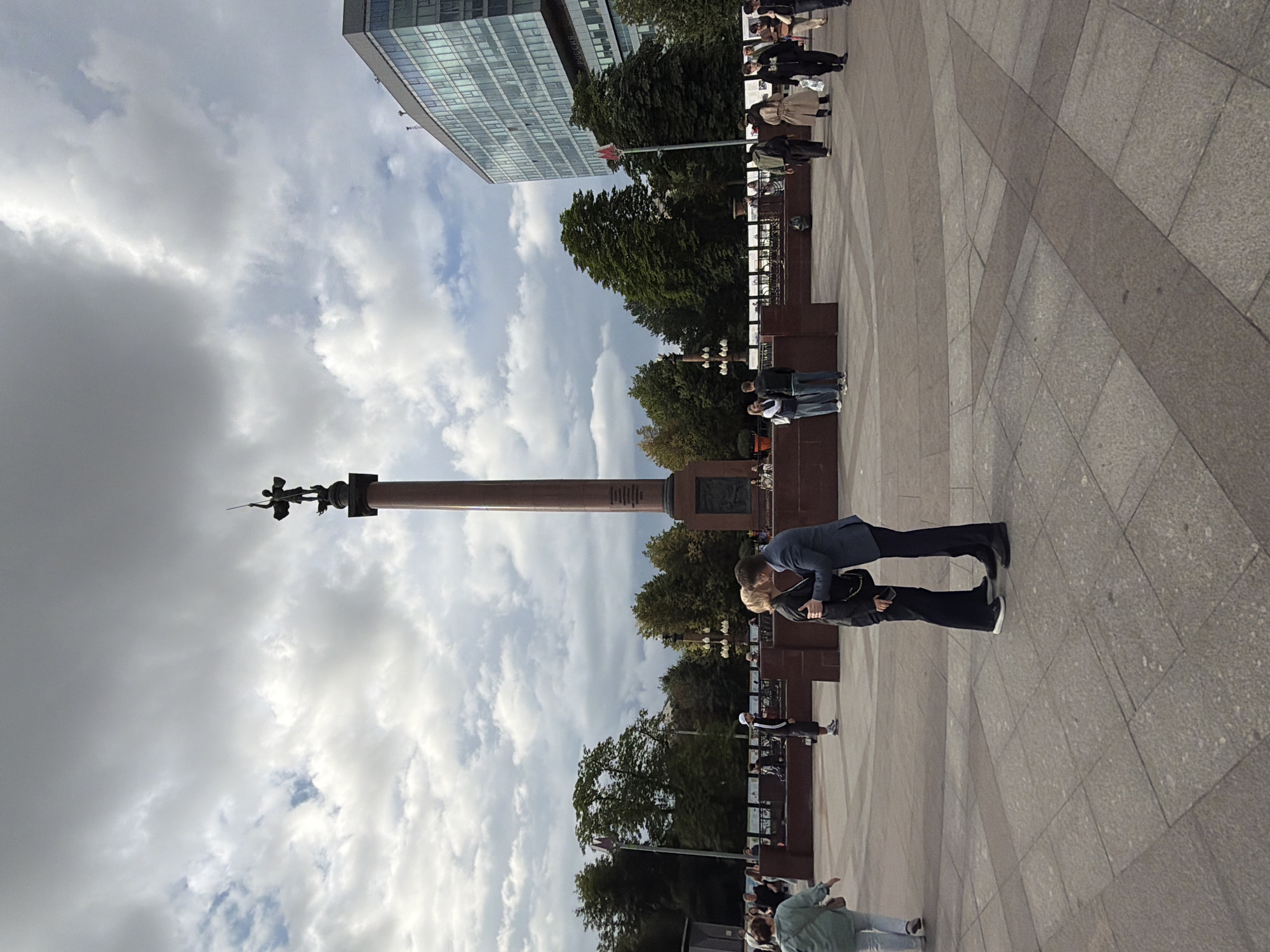 The image depicts an outdoor scene with a tall, reddish-brown monument topped with a statue of a winged figure holding a spear. The monument stands against a backdrop of a cloudy sky with patches of blue. To the left of the monument is a modern glass-fronted building. In the foreground, on a paved plaza, several people are visible. A couple is in the process of kissing, with one person bending down to meet the other. Other individuals are walking or standing around, some appearing to be tourists. The paving stones of the plaza are laid in a geometric pattern. Trees are visible behind the monument. The time of day appears to be daytime, and the weather is overcast with some sunshine breaking through the clouds. No discernible text is visible in the image.