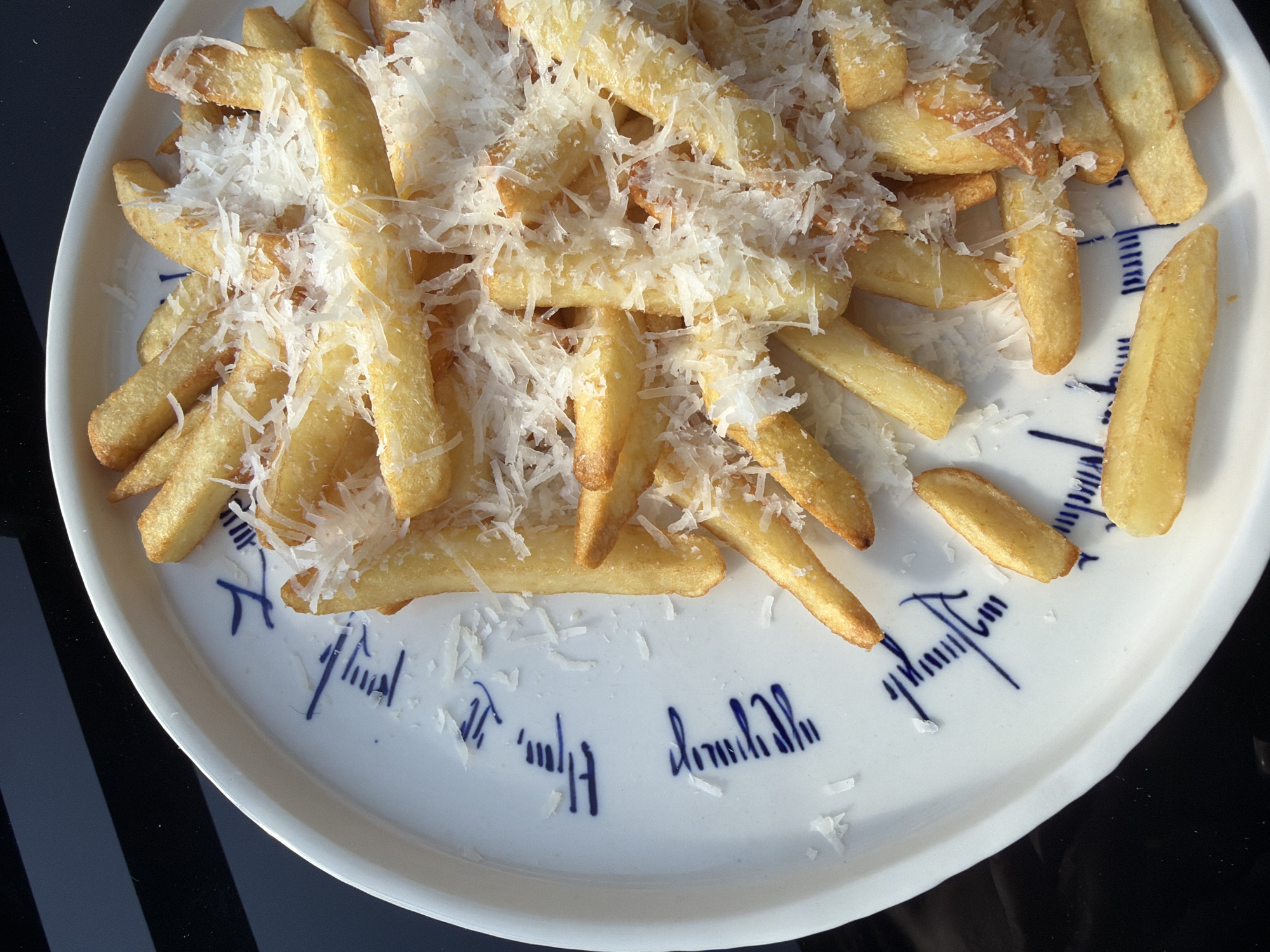 A high-angle shot reveals a white, round plate filled with French fries. The fries are topped with shredded cheese. The plate features a decorative blue pattern. The plate sits on a dark, reflective surface, likely a table. The location is unknown and cannot be determined from the image.