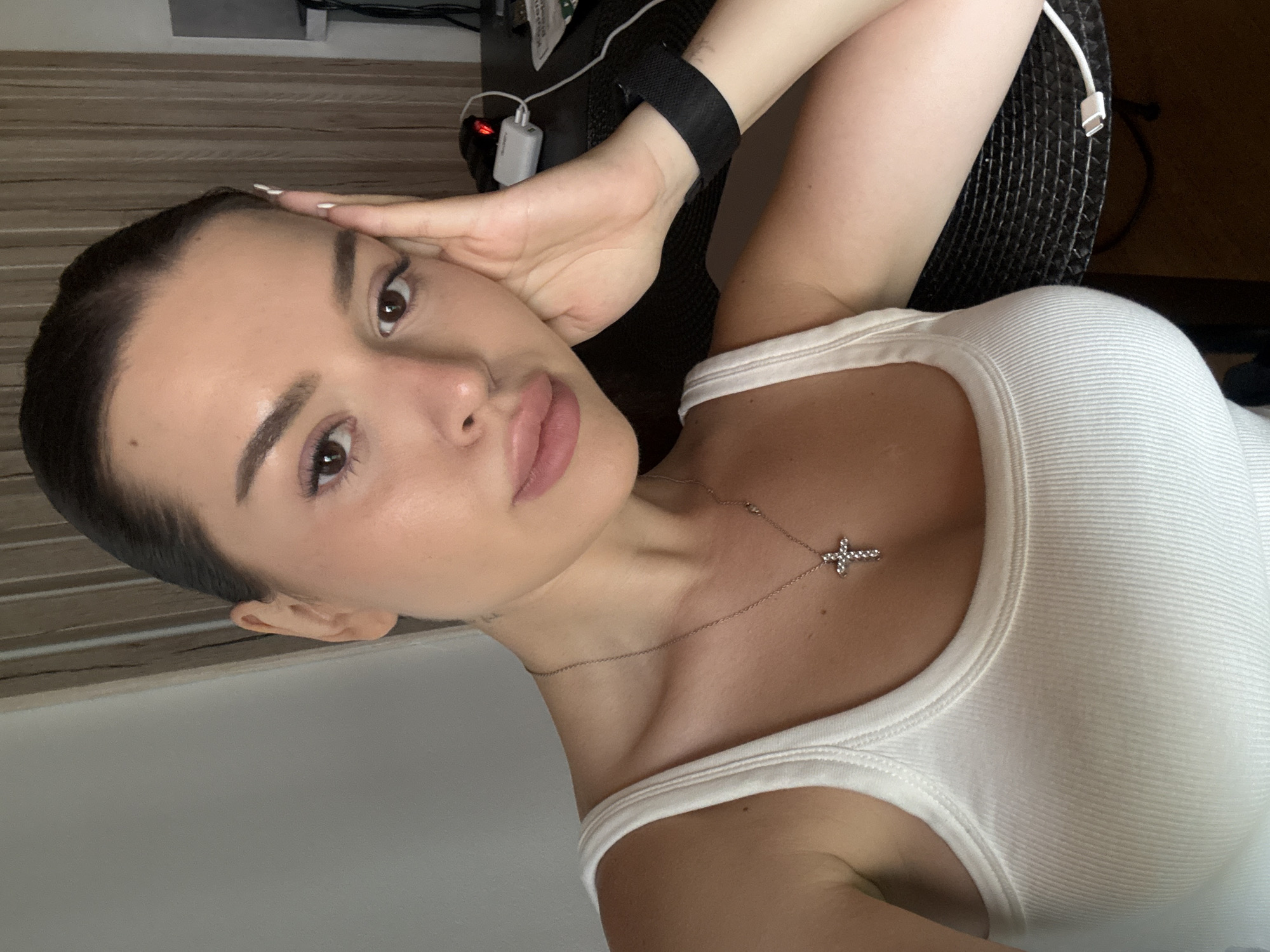 A young woman with dark hair pulled back, wearing a white ribbed tank top and a silver cross necklace, is positioned in the foreground of the image. She is looking directly at the camera, with her right hand resting on her forehead and her left arm visible. Her facial features include defined eyebrows, brown eyes, and full lips. The background suggests an indoor setting, possibly a room, with vertical blinds on the right side and a wall on the left. A black object, possibly a desk or table, is partially visible, with a white charging cable and a small white device on it. The lighting appears to be natural or from an overhead source, casting soft shadows. The image's resolution is clear, allowing for detailed observation of the subject and her immediate surroundings. The location is not identifiable as Prague, Czechia.