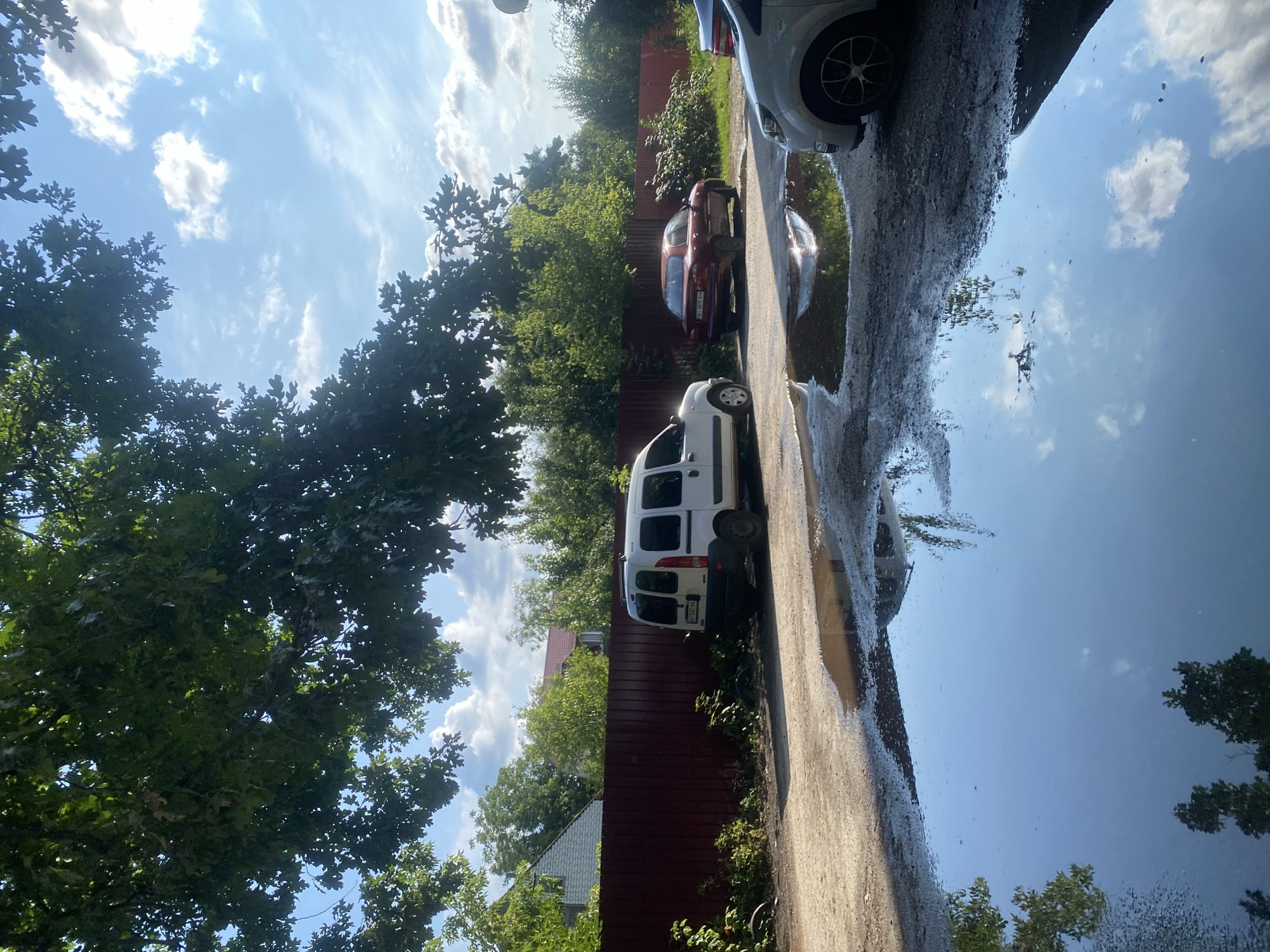 The image depicts a sunlit outdoor scene with a large puddle in the foreground, reflecting the sky, clouds, and surrounding trees. Beyond the puddle, a gravelly area leads to a red wooden fence. A white minivan is parked to the right of the fence, and a dark red sedan is parked further down. In the background, houses and dense green trees are visible under a bright blue sky with scattered white clouds. The scene appears to be a residential area or a parking lot. Based on visual cues, this could be a scene from Poltava, Ukraine, given the architectural styles of the houses and the types of vehicles present.