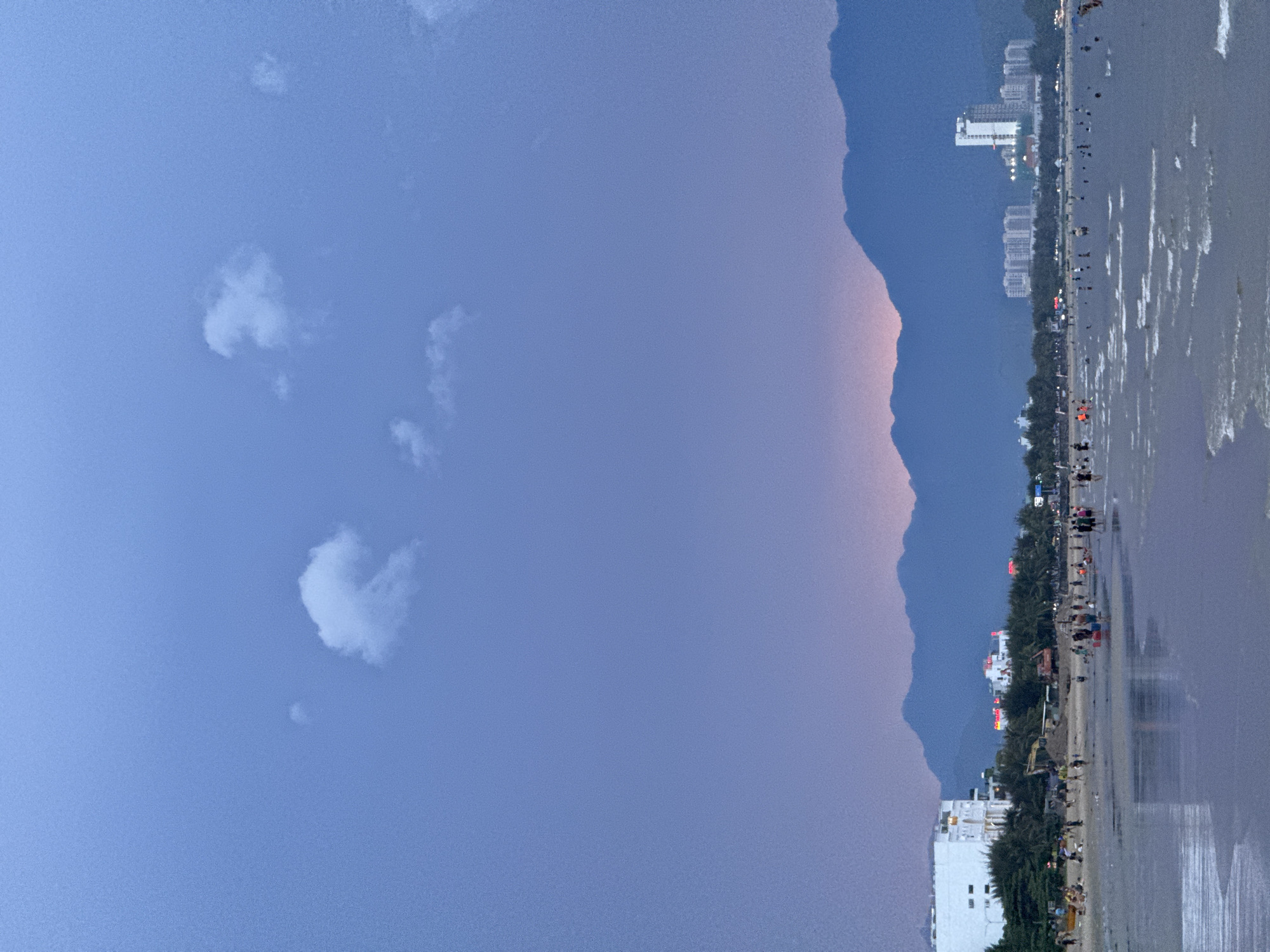 The image depicts a beach scene at dusk in Đà Nẵng, Vietnam. The sky is a gradient of deep blue to soft purple, with a few scattered white clouds. The calm ocean reflects the twilight sky, with gentle waves lapping at the shore. Along the coastline, a row of palm trees separates the beach from a developed area featuring several white, multi-story buildings and a road. Numerous people are visible walking along the beach and the promenade, some appearing to be tourists or locals enjoying the evening. The overall atmosphere is peaceful and serene. The time of day is evident from the soft, ambient light and the purple hues in the sky, suggesting sunset or shortly after. The location cues, such as the architecture of the buildings and the presence of palm trees, are consistent with a coastal city in Southeast Asia. There are no discernible objects or activities that indicate a specific event. The mood is calm and relaxed. Some neon signs with Vietnamese text are visible on the buildings, but the text is not legible.