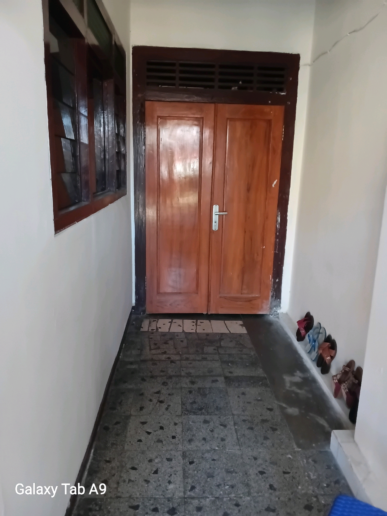 This is a photograph of an indoor hallway leading to a double wooden door. The hallway has a tiled floor with dark gray and black speckled tiles. To the left, a white wall meets a window frame with dark wood slats. To the right of the door, several pairs of sandals are lined up against the white wall, including red, blue, brown, and dark patterned pairs. Above the door is a dark wooden ventilation grate. The photograph is taken from a slightly low angle, and the lighting suggests it is daytime. The text "Galaxy Tab A9" is watermarked in the lower left corner. The location is identified as Kelutan, Indonesia.