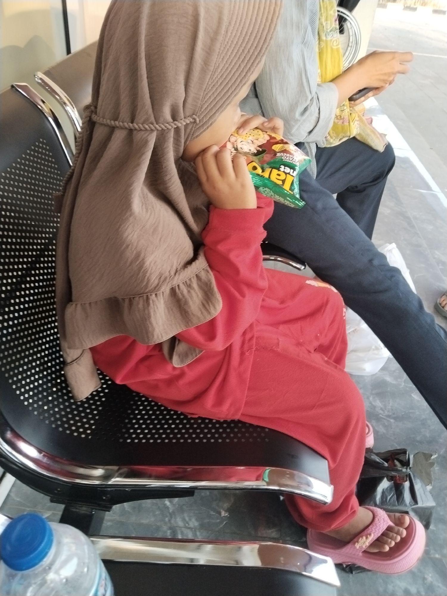 A young child wearing a brown hijab and a red outfit is seated on a black perforated chair. The child is eating snacks from a green and yellow bag. To the right of the child, an adult is seated, wearing a light blue shirt and dark pants. The adult is holding a mobile phone and has a yellow cloth draped over their left shoulder. A plastic water bottle with a blue cap is visible in the foreground on the left. The scene appears to be an indoor waiting area, possibly at a public facility in Sentul, Indonesia, indicated by the style of seating and the general environment.