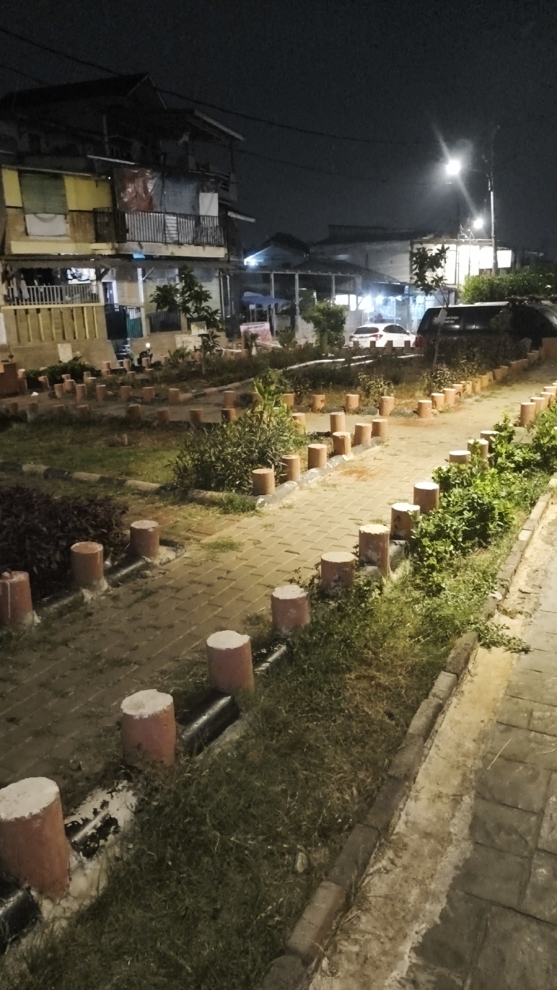 The image depicts an outdoor scene at night, likely in a residential or public area, with artificial lighting illuminating the surroundings. In the background, there are multi-story buildings that appear aged, with some structures showing signs of wear and tear, including visible tarps and exposed materials. A street lamp is visible, casting a bright light.
In the mid-ground, a paved pathway curves through a landscaped area. The pathway is lined with small, cylindrical posts, some appearing to be made of concrete with reddish-brown exteriors and white tops. There are also patches of grass and some low-lying shrubs and plants. Several parked vehicles are visible in the distance, including a dark-colored SUV and a white car.
The foreground shows a section of the paved pathway and the grass verge. The pathway is made of interlocking bricks. The grass verge contains more of the cylindrical posts and some darker, denser foliage. The overall impression is of an urban environment at night, with an attempt at landscaping that appears somewhat unkempt.
This scene is likely located in a densely populated urban area of South Jakarta, Indonesia, given the architectural style of the buildings and the common urban landscaping features.