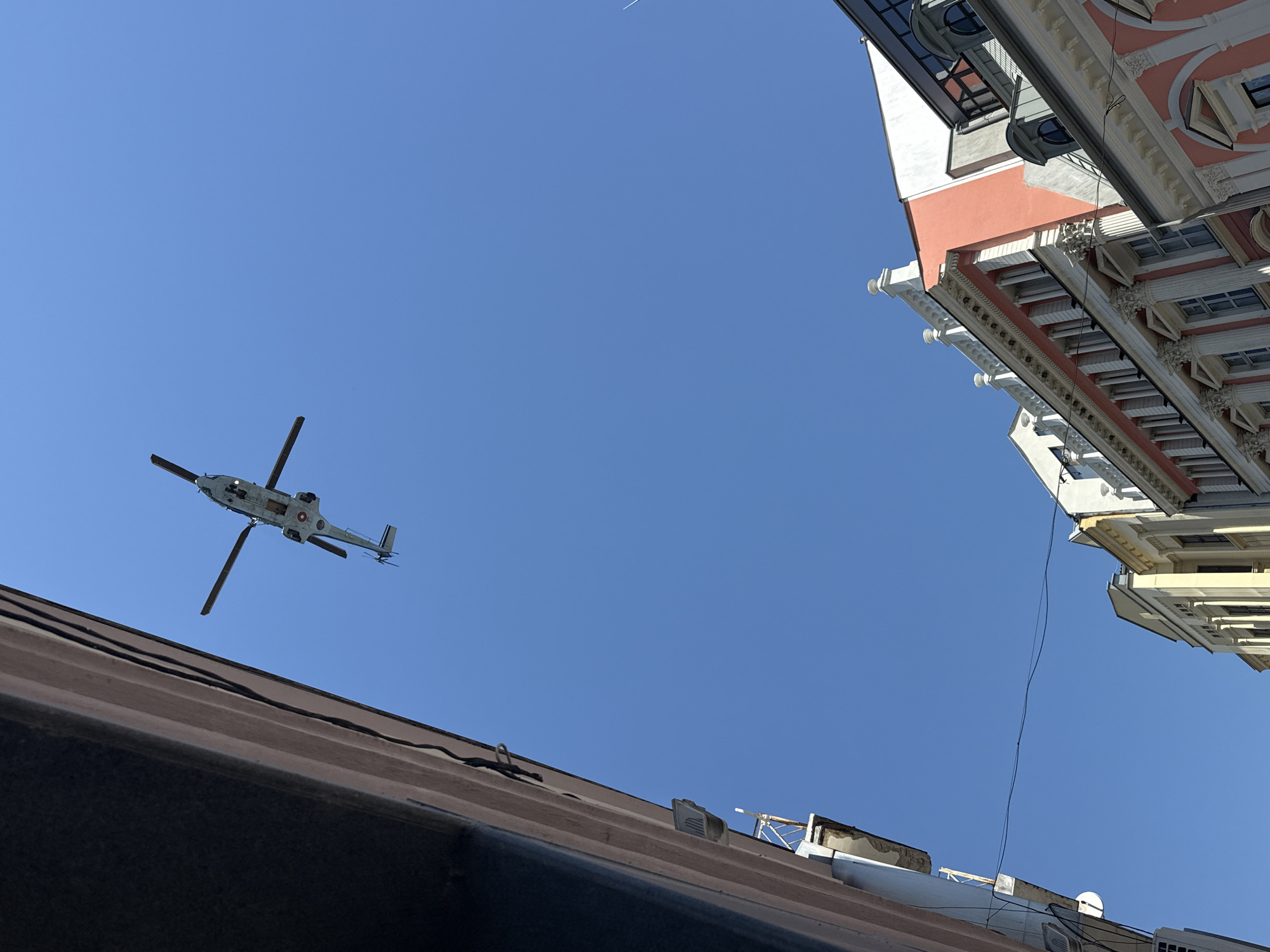 The image is a low-angle shot capturing a helicopter in flight against a clear blue sky, juxtaposed with architectural elements.
In the lower foreground, there is a dark, sloped roof with visible seams and rooftop fixtures. A white rectangular object and a small antenna are also present on the roof.
Above the roof, the sky is a solid blue, against which a light-colored helicopter is visible. The helicopter's rotor blades are in motion. The helicopter's shape and markings are visible.
To the right of the sky, the upper corner of a tall, ornamented building intrudes into the frame. The building's facade is a mix of colors, including cream, peach, and white, with ornate details like window frames, cornices, and decorative elements. A black cable also droops in front of the building.
Based on the architecture and available information, this image was likely taken in Plovdiv, Bulgaria, where such historical buildings and air traffic are found.