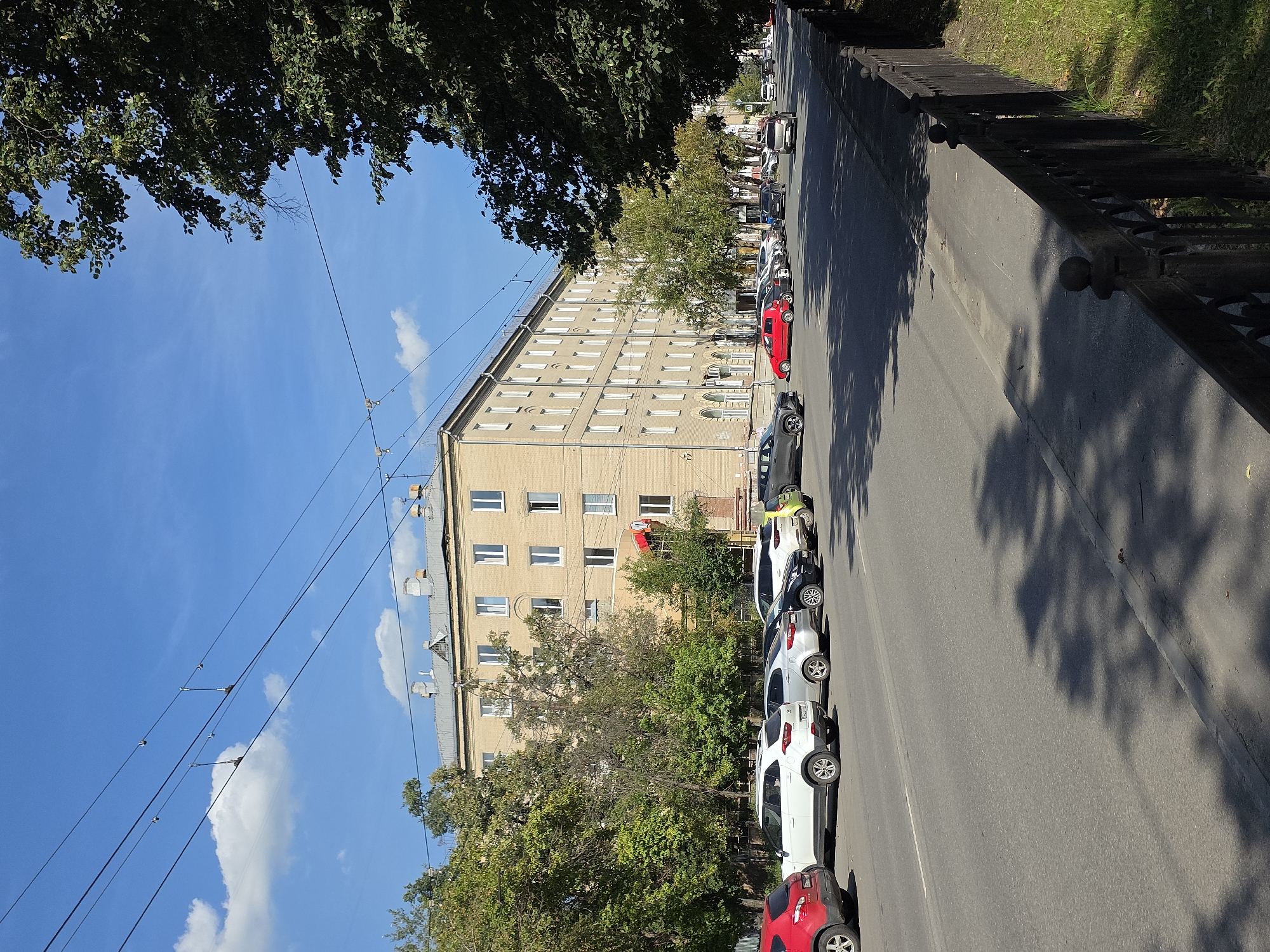 The image depicts a street scene in Chelyabinsk, Russia, on a sunny day. A long, light-yellow apartment building with numerous white-framed windows dominates the left side of the frame. Trees line the sidewalk in front of the building, casting shadows on the street. Several cars are parked along the curb on the right side of the street, including a red hatchback, a white SUV, and other vehicles. The street itself is asphalt, with a white line marking the lane. Overhead, power lines crisscross the bright blue sky, with a few white clouds visible. On the far right, a dark metal fence separates the sidewalk from a grassy area. The overall impression is of a typical urban residential street.