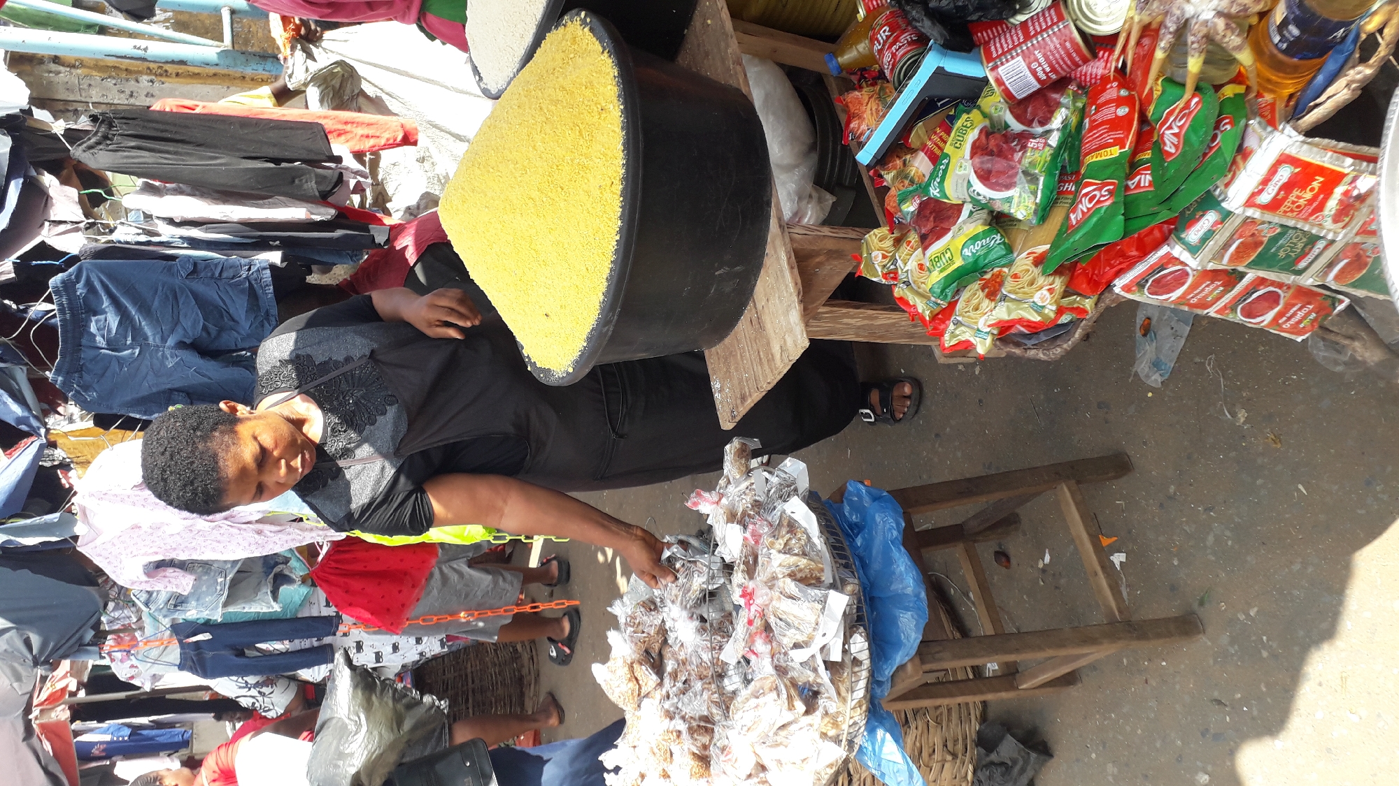 A market scene in Onitsha, Nigeria shows a woman seated among clothing items, groceries and a black bucket filled with yellow grains. She has short black hair, is wearing a black patterned top and black pants. A wooden table holds red and green packets of tomato paste and noodles and cans, while a smaller, wooden table holds dried fish in clear plastic packaging. She reaches toward the dried fish. Clothing hangs on a rack behind her. Shoppers' feet are visible around the tables.