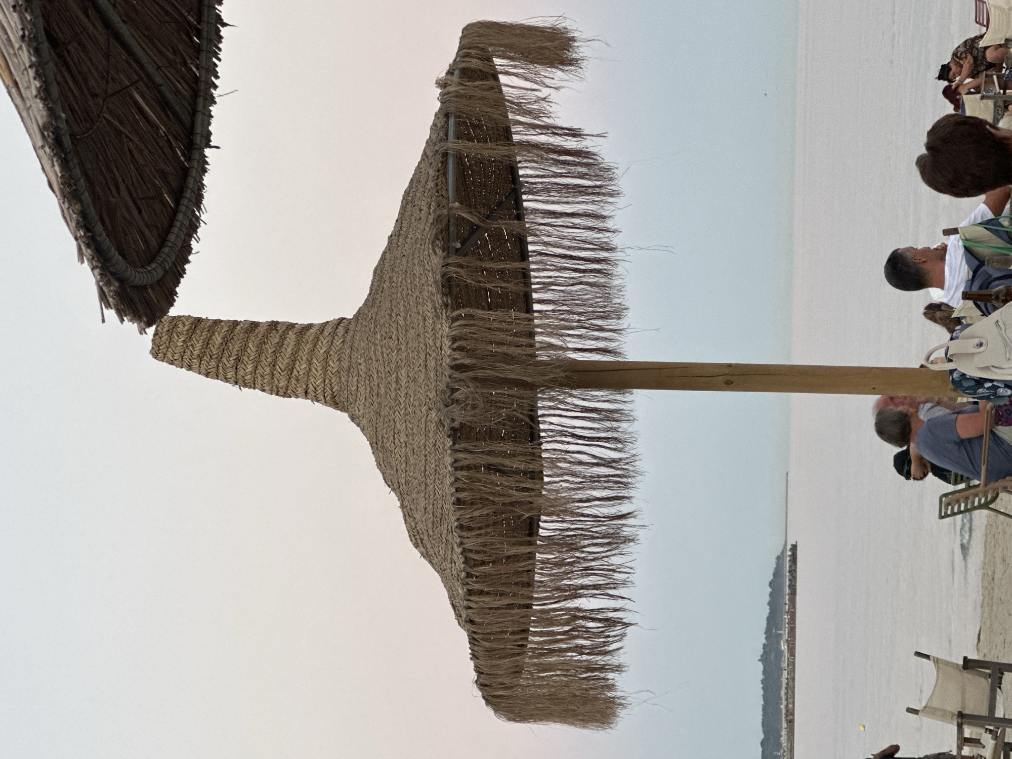 The image depicts a tranquil scene at a beachside establishment, likely a cafe or restaurant, in Varna, Bulgaria. Two prominent thatched umbrellas, characteristic of seaside venues, dominate the foreground. The larger umbrella is a focal point, featuring a conical top and a wide brim adorned with a fringe of natural fibers. A wooden pole supports this umbrella. Beneath the umbrella, several patrons are seated on chairs, some facing the sea, while others are engaged in conversation. The sea stretches out in the background, calm and reflecting the overcast sky. In the distance, a breakwater or pier is visible, with buildings lining its edge. The overall atmosphere is relaxed and unhurried, suggesting a leisurely afternoon.
