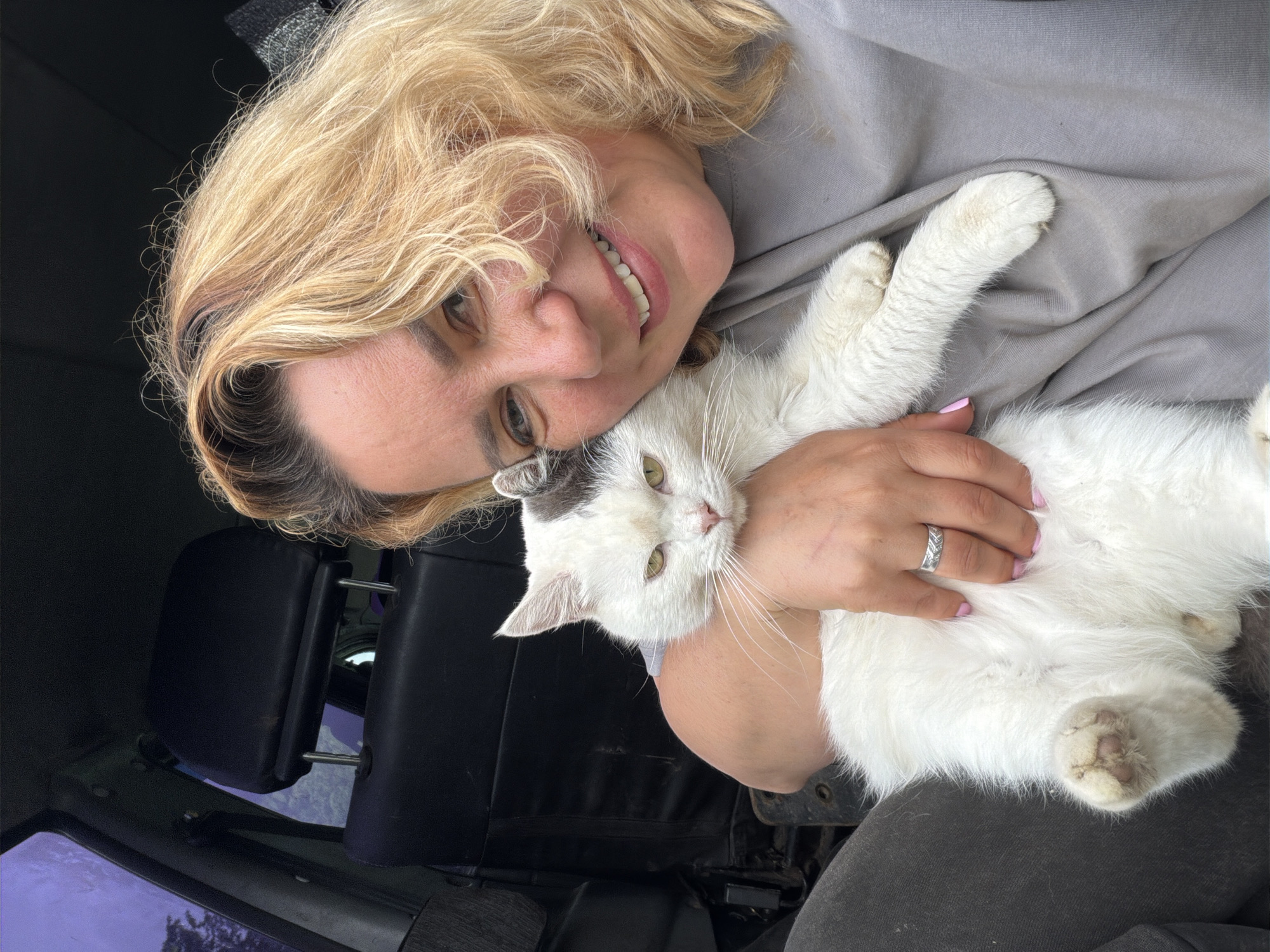 A woman with blonde hair is smiling and holding a white cat with gray markings in her arms. She is wearing a gray shirt and has a silver ring on her left ring finger. The cat has green eyes and long white whiskers. The background appears to be the interior of a vehicle with black seats. The location is Leningradskaya, Russia.