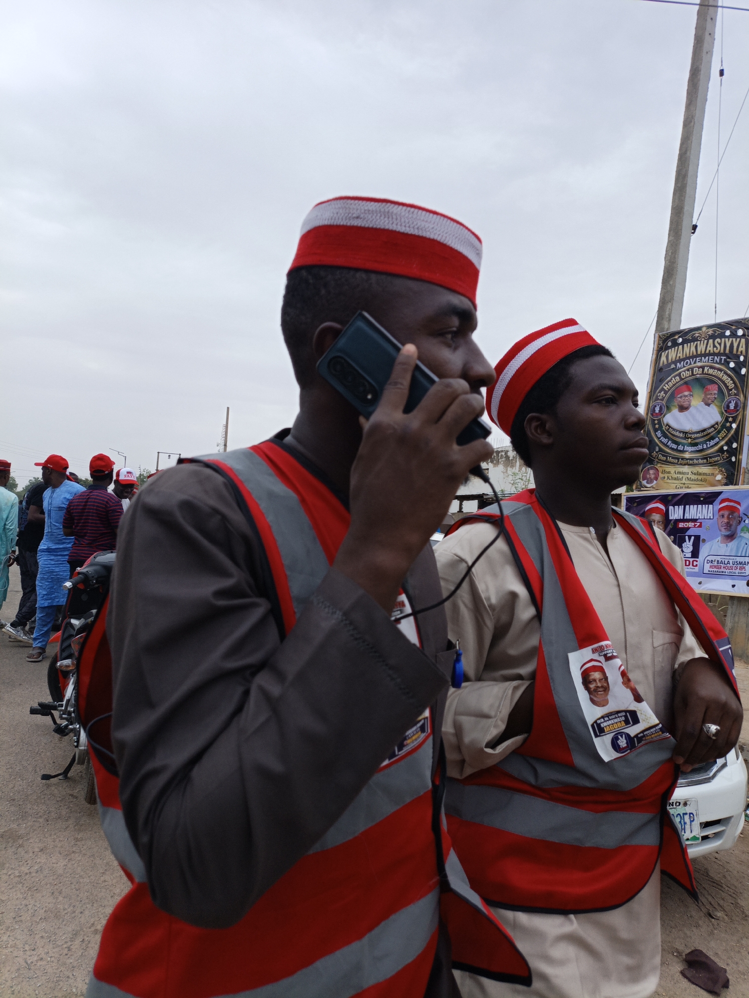 The image shows two men wearing red and white hats and reflective vests. One man is on a phone call, while the other looks off into the distance. They appear to be at an event or rally, as indicated by the political banners in the background. One banner says "KWANKWASIYYA MOVEMENT" and features images of political figures. Other people are visible in the background, some also wearing red hats. A motorcycle is parked nearby. The license plate visible in the foreground indicates location in Nigeria.
